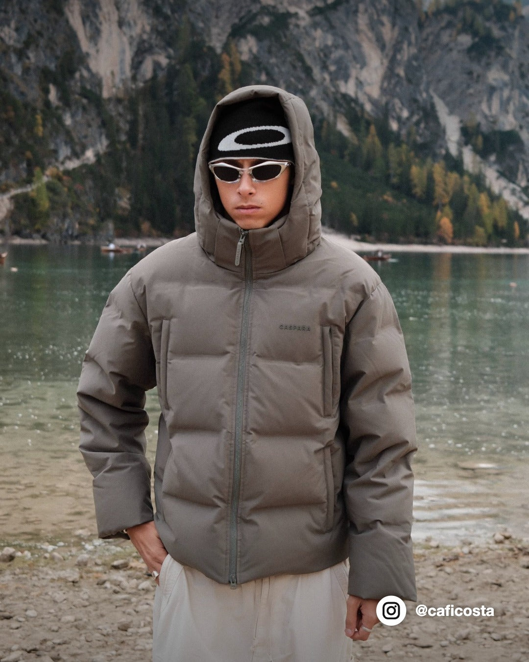 Man wearing a khaki CASPARA Boreal puffer jacket standing in front of a mountain lake.