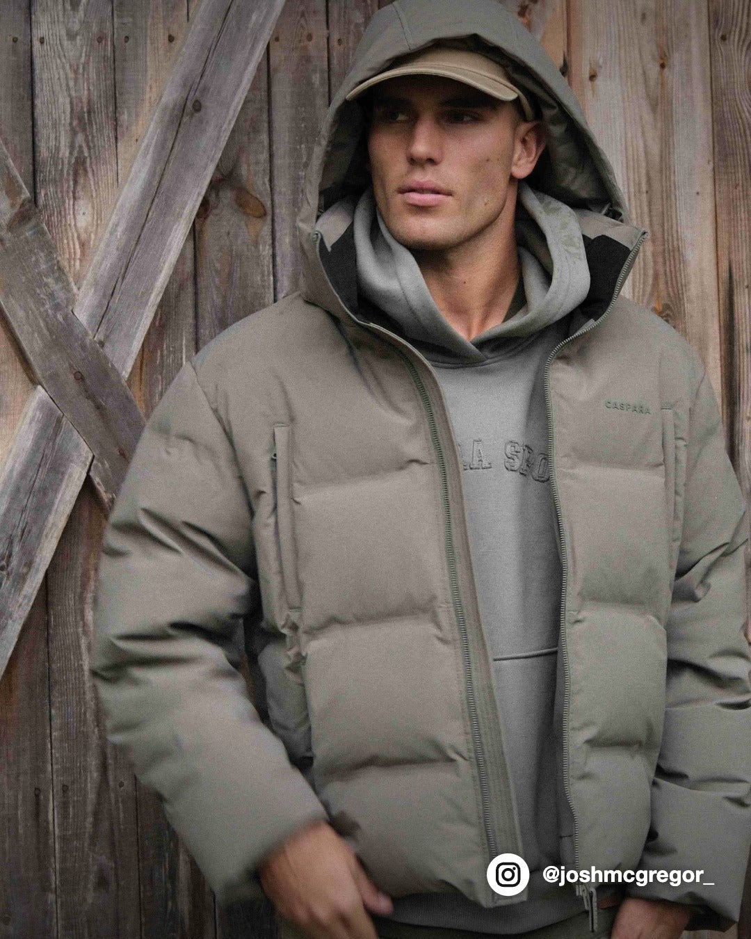 Man wearing a khaki CASPARA Boreal puffer jacket with hood up against a wooden wall.