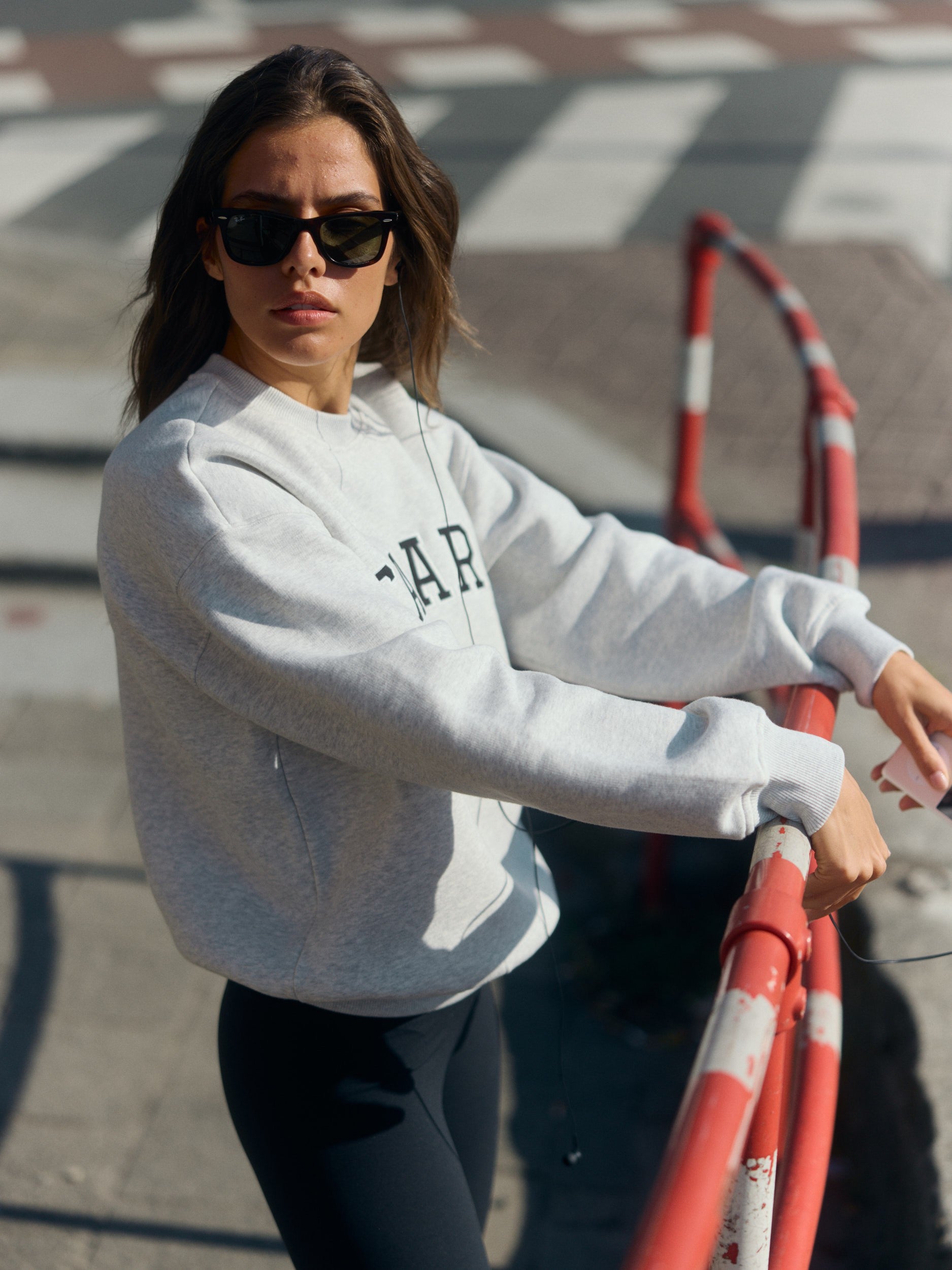 Woman wearing a grey melange crewneck sweatshirt and sunglasses leaning on a street barrier.