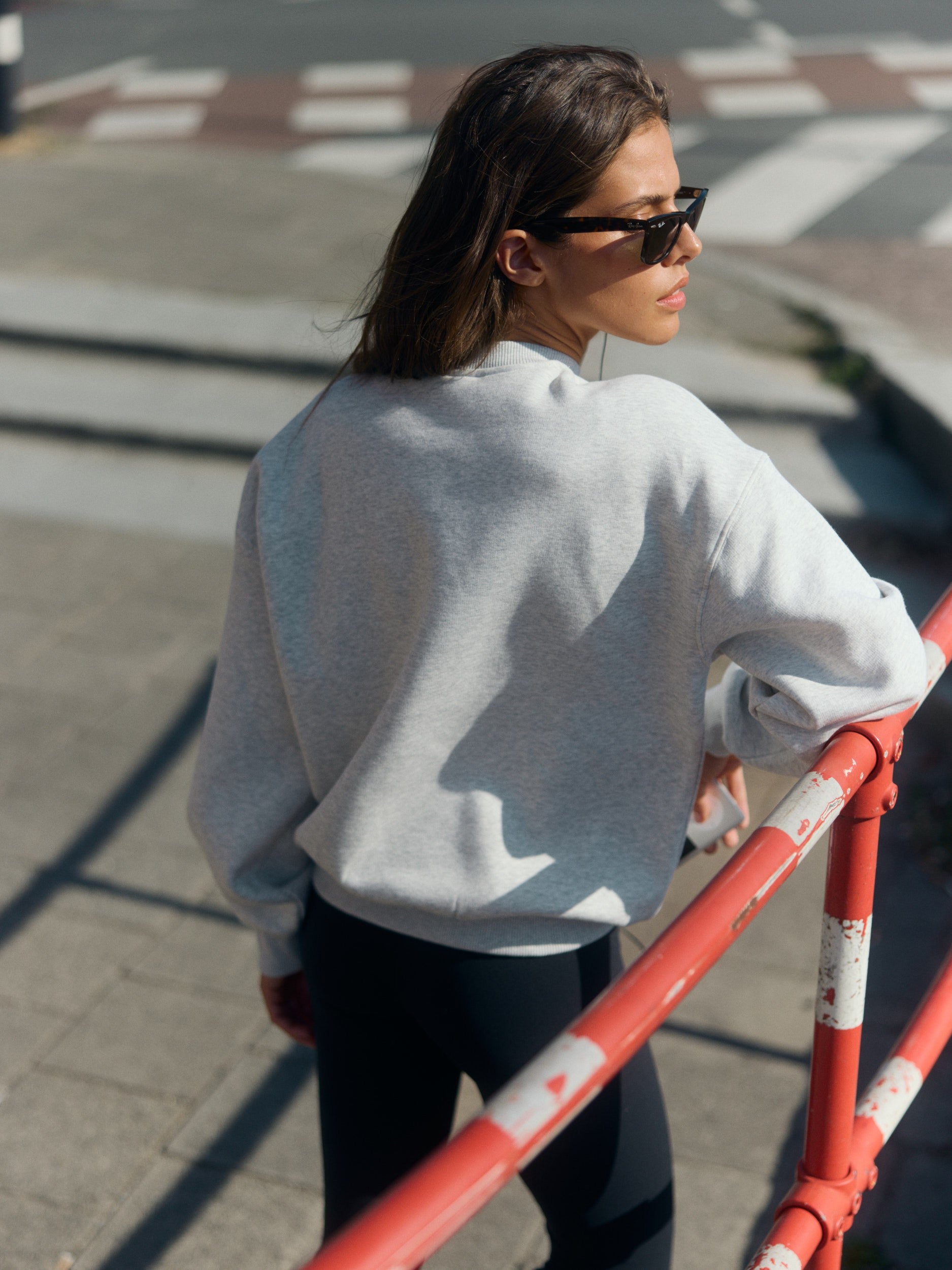 Woman wearing a grey melange crewneck sweatshirt leaning against a red railing outdoors.