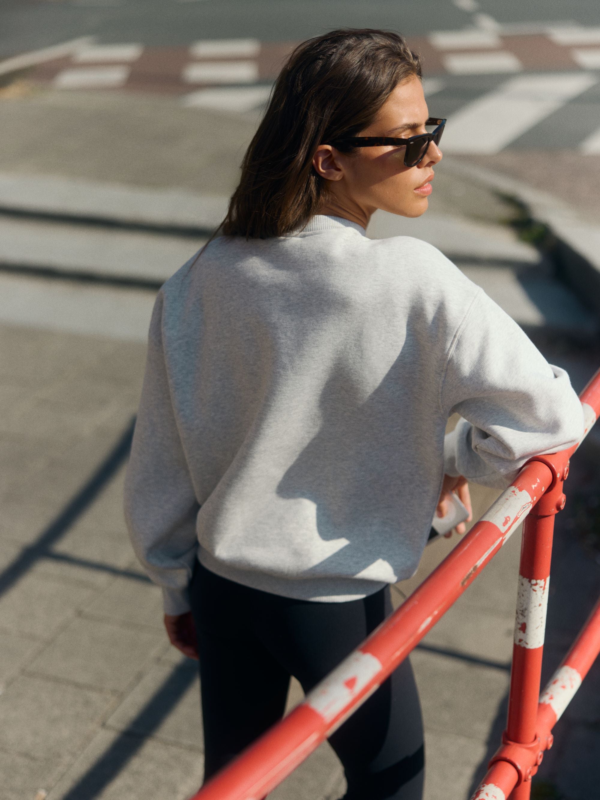Woman wearing a grey melange crewneck sweatshirt and sunglasses leaning on an outdoor railing.
