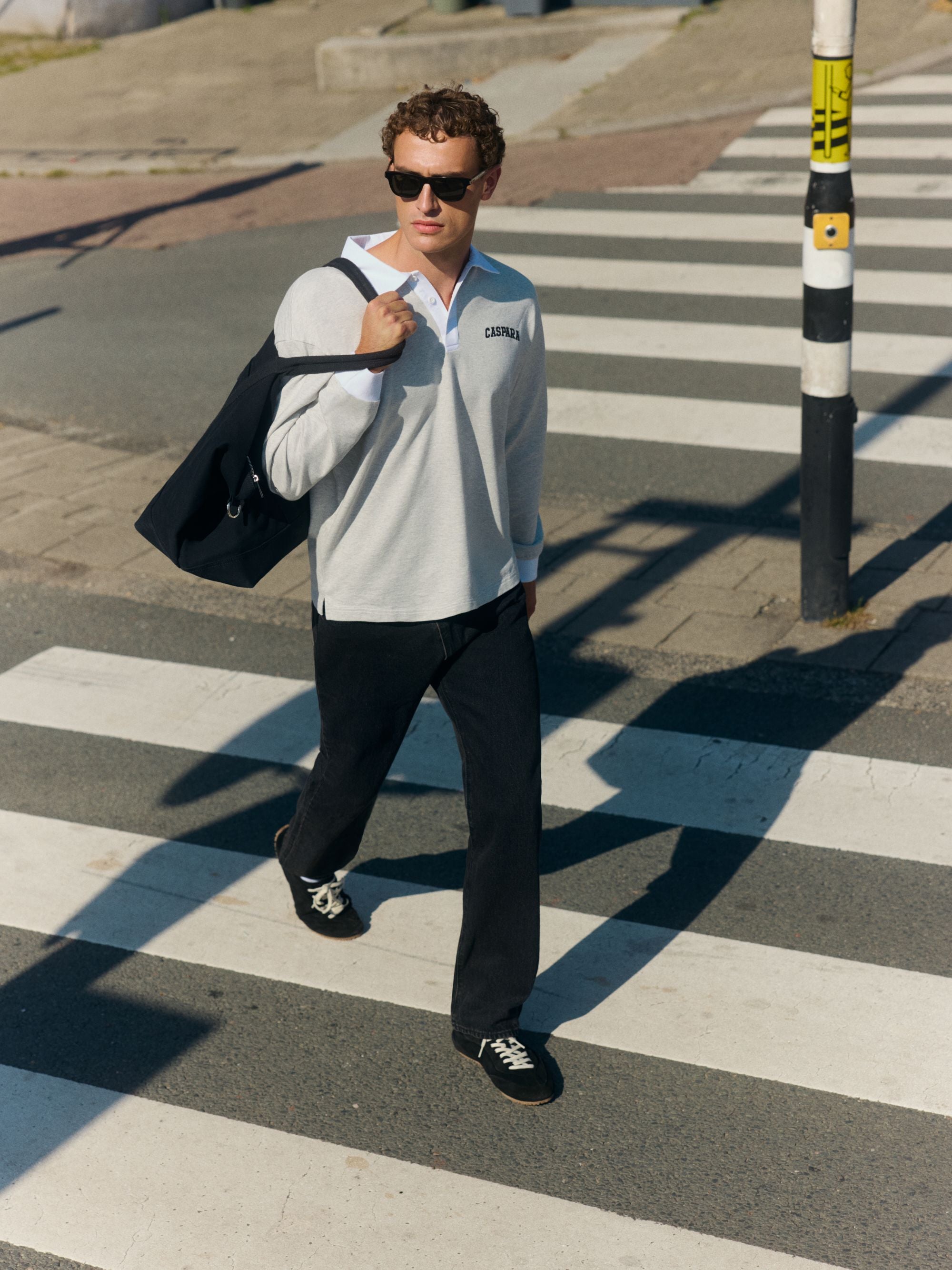 Man wearing a grey and white CASPARA rugby shirt and sunglasses walking across a crosswalk.