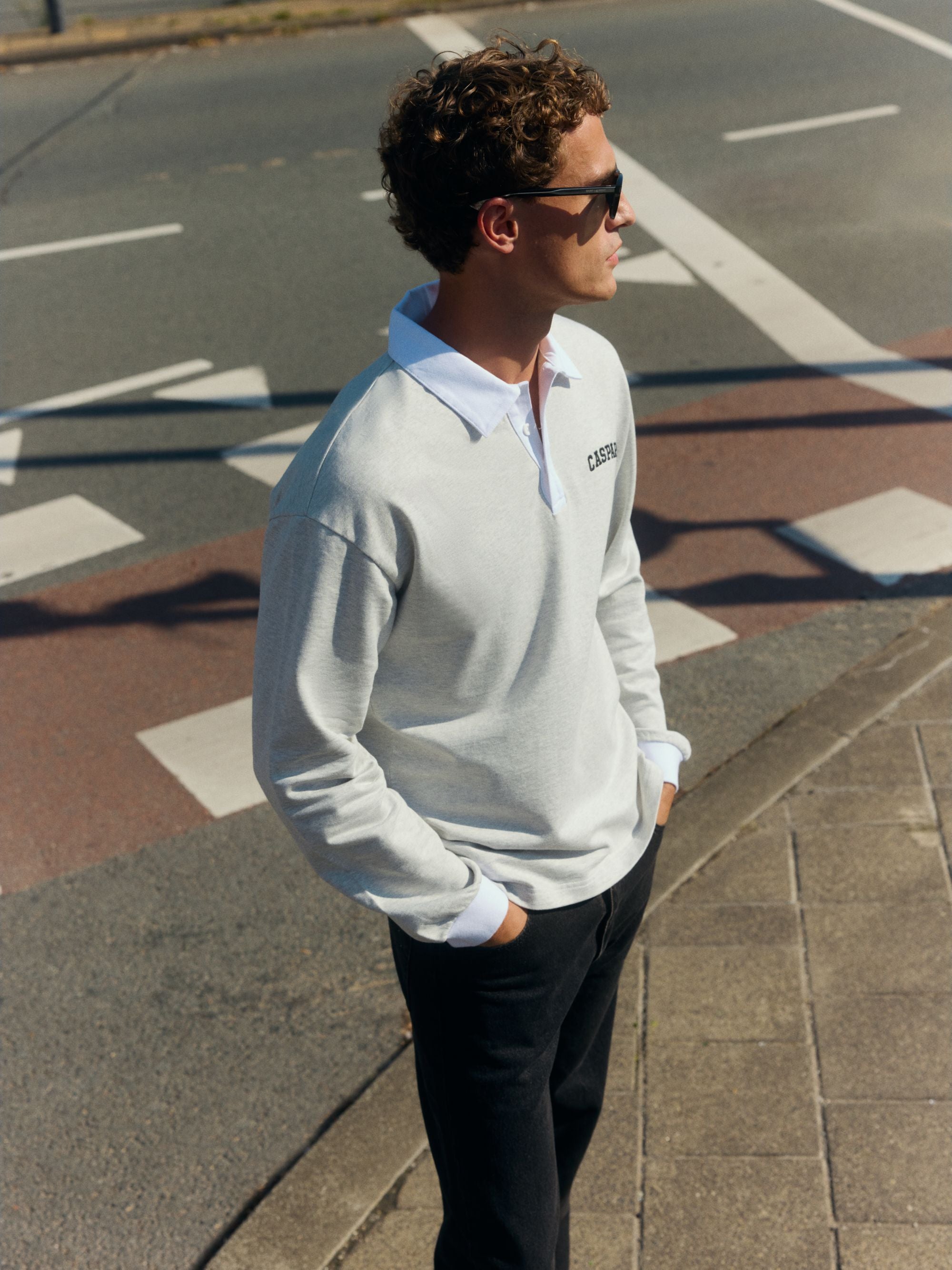 Man wearing a grey and white CASPARA logo rugby shirt and sunglasses on a city street.
