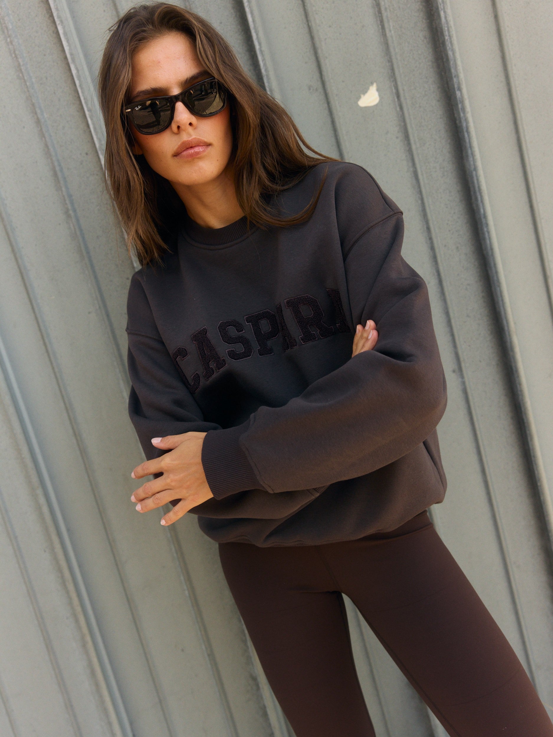 Woman wearing an espresso brown oversized crewneck sweatshirt and sunglasses against a metal wall.