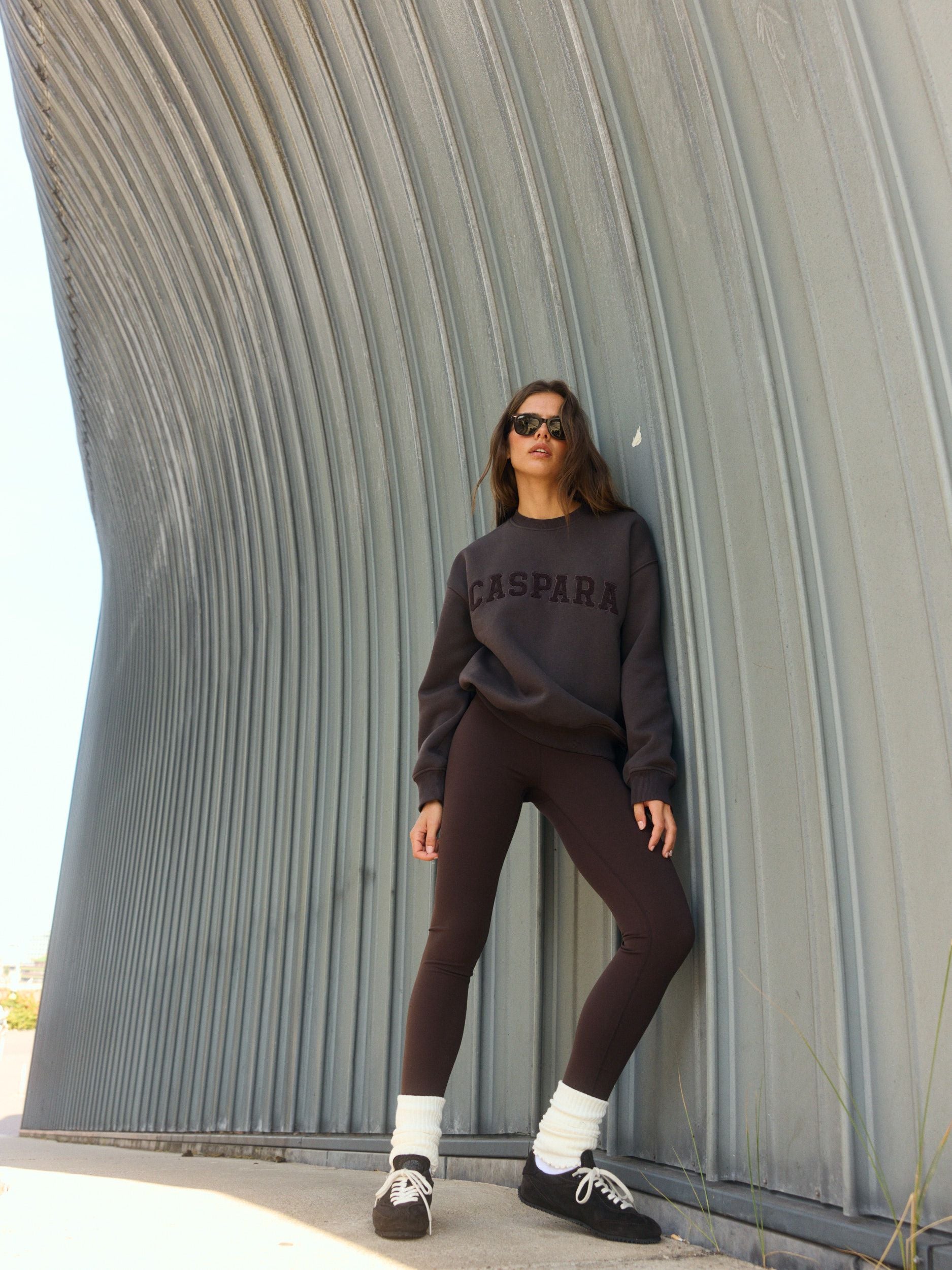 Woman wearing an espresso brown CASPARA crewneck sweatshirt and matching leggings against a curved metal wall.