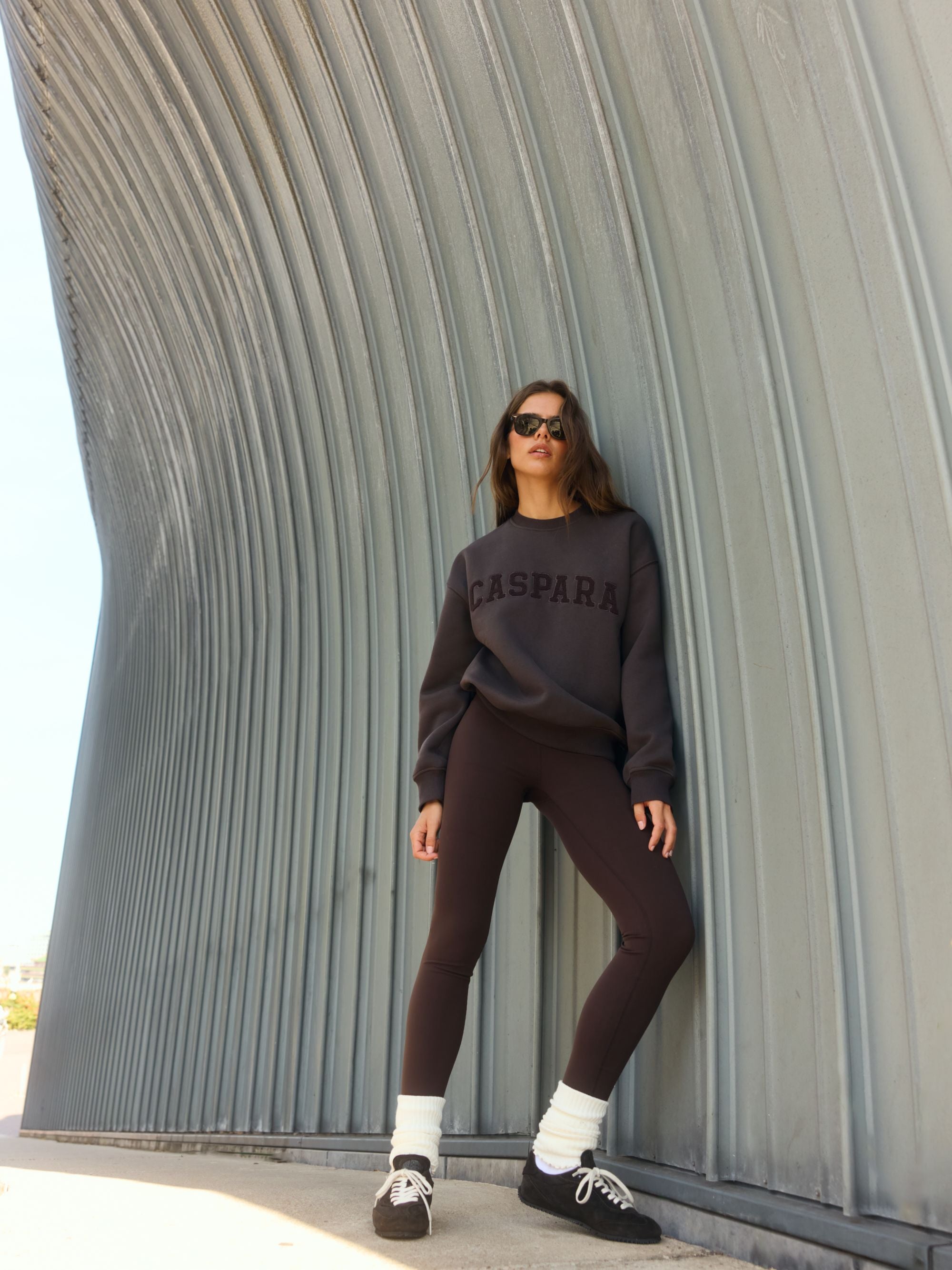 Woman wearing an espresso brown CASPARA logo crewneck sweatshirt and sunglasses outdoors.