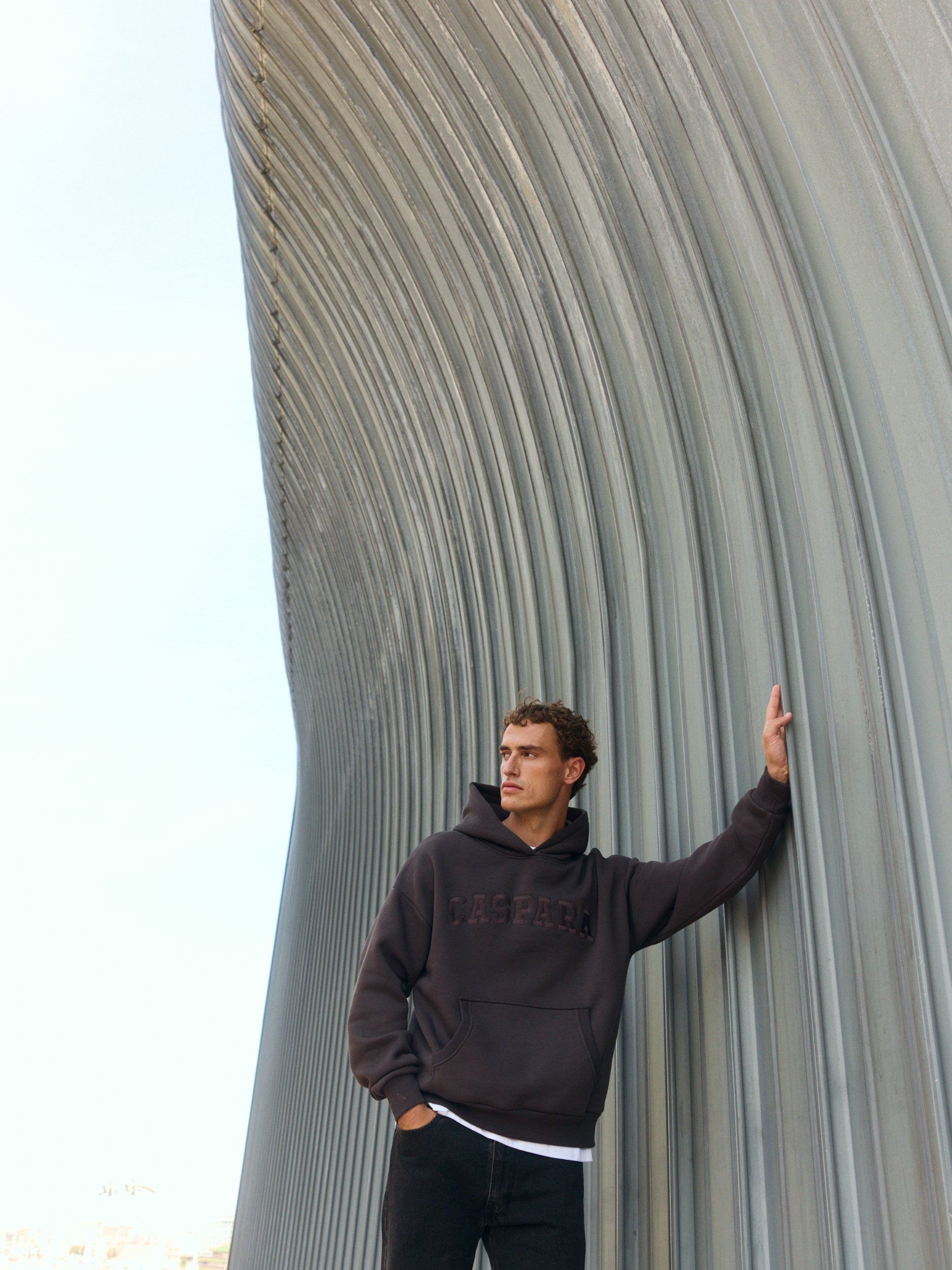 Man wearing an espresso brown hoodie leaning against a curved architectural wall.