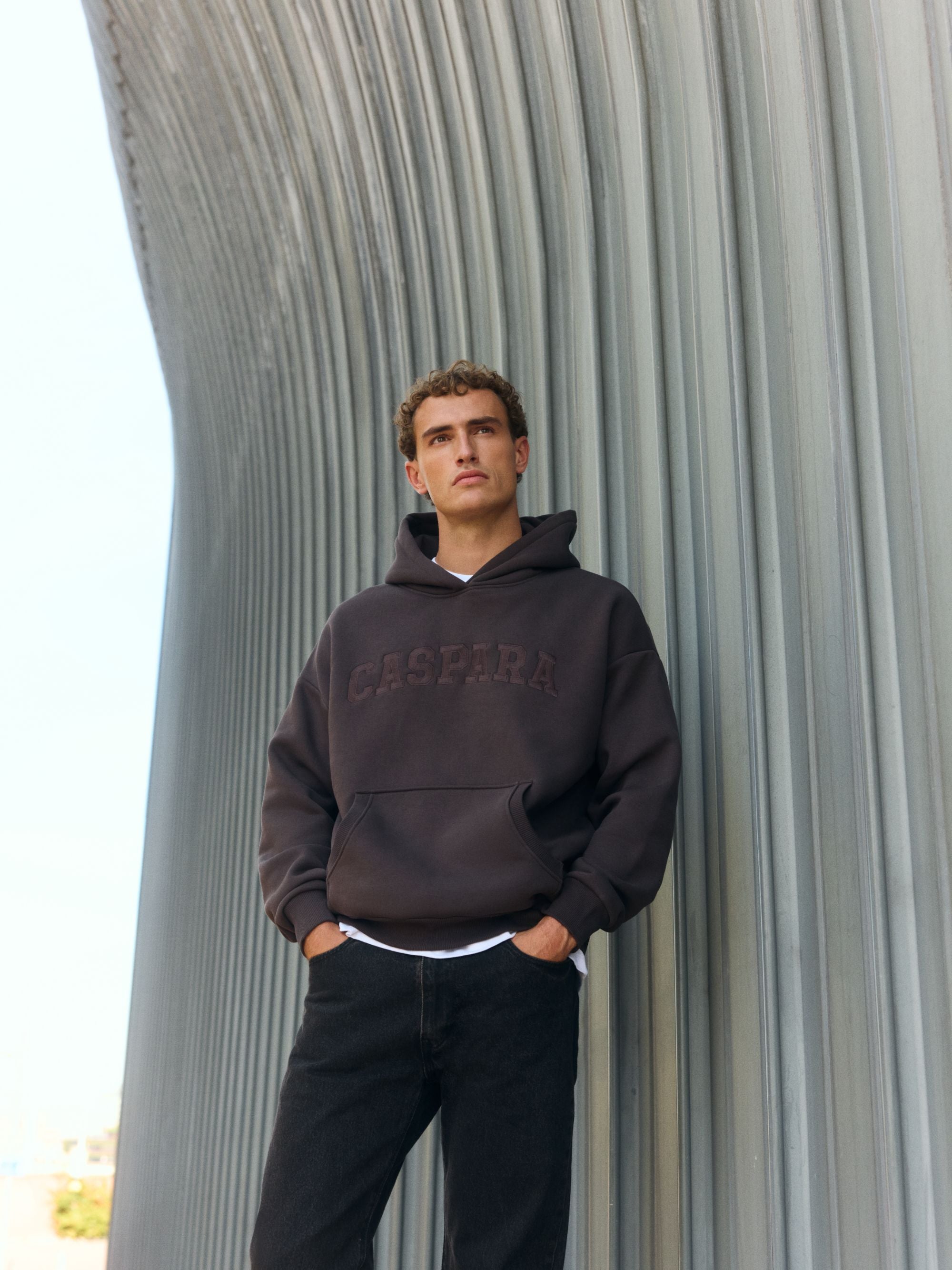 Man wearing an espresso CASPARA logo hoodie in front of a modern architectural wall.