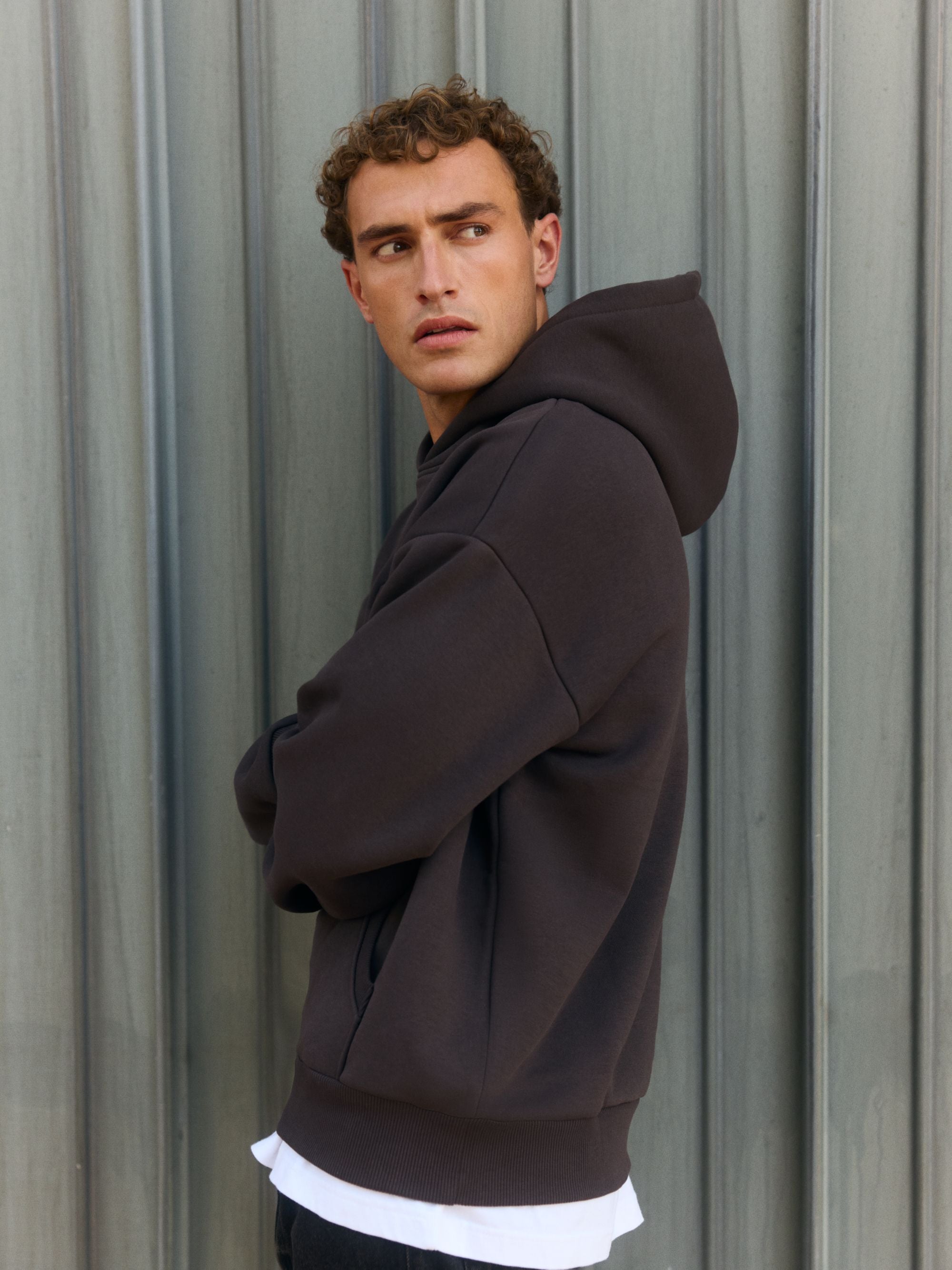 Man wearing an espresso CASPARA All-Star hoodie against a corrugated metal wall.