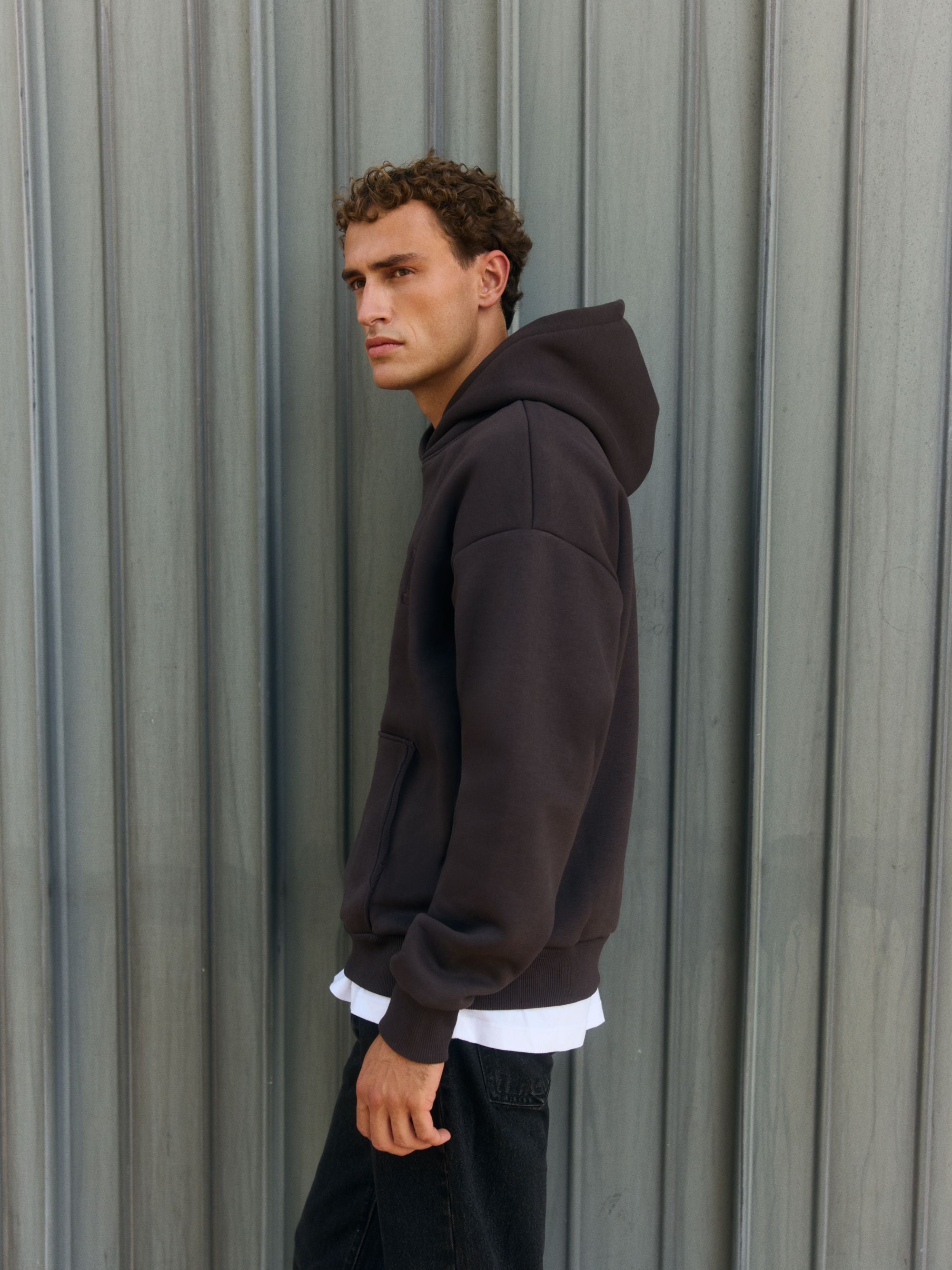 Man wearing an espresso CASPARA All-Star hoodie against a grey corrugated wall.