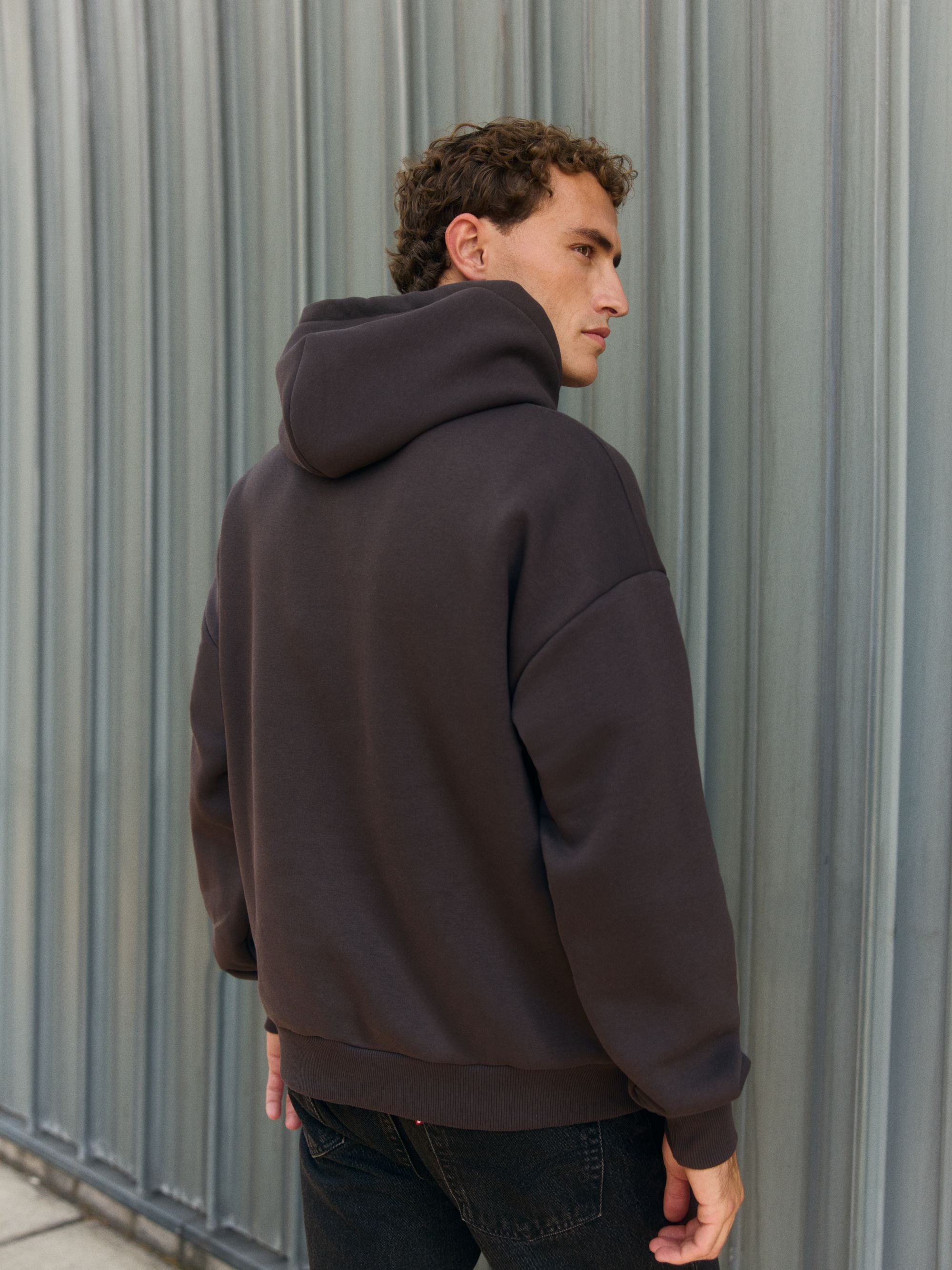Back view of a man wearing an espresso brown CASPARA hoodie against a grey metal wall.