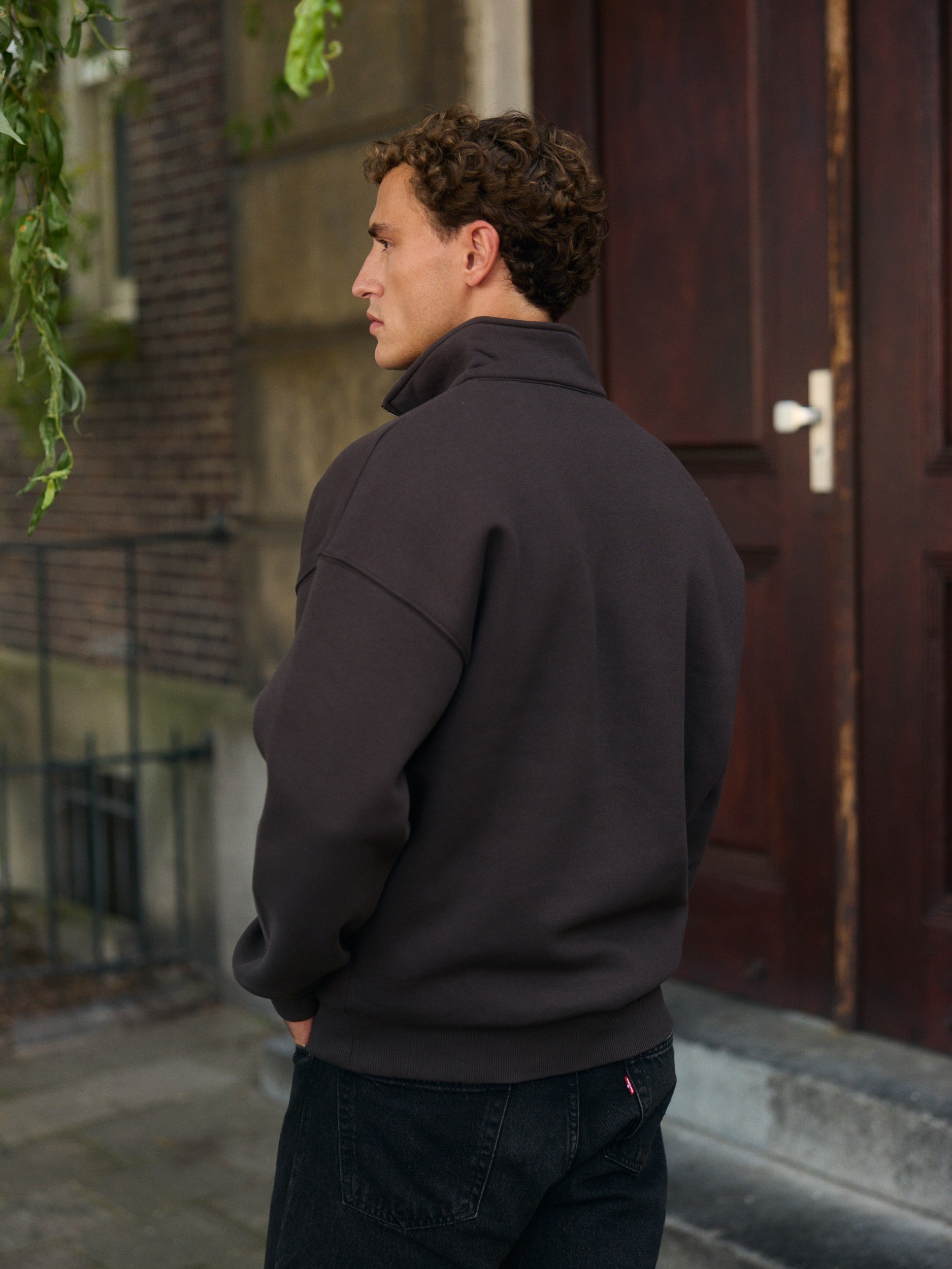 Man wearing an espresso brown quarter-zip sweatshirt standing outside in front of a wooden door.