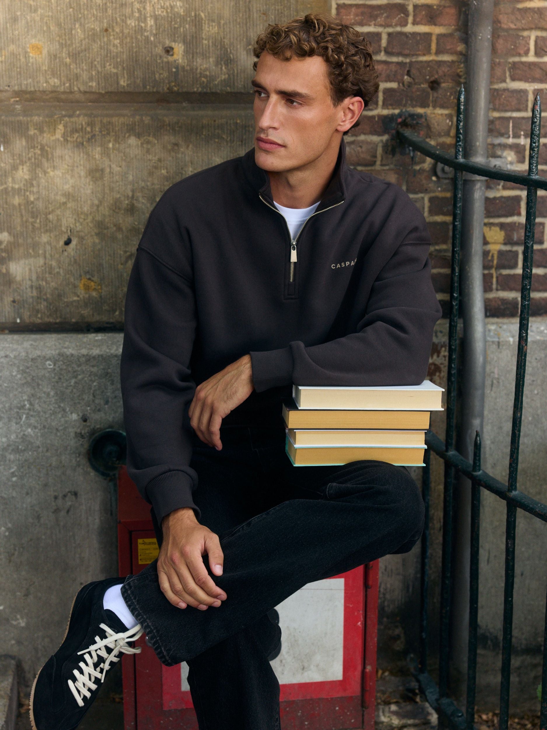 Man wearing an espresso brown quarter-zip sweatshirt leaning on a stack of books against a brick wall.