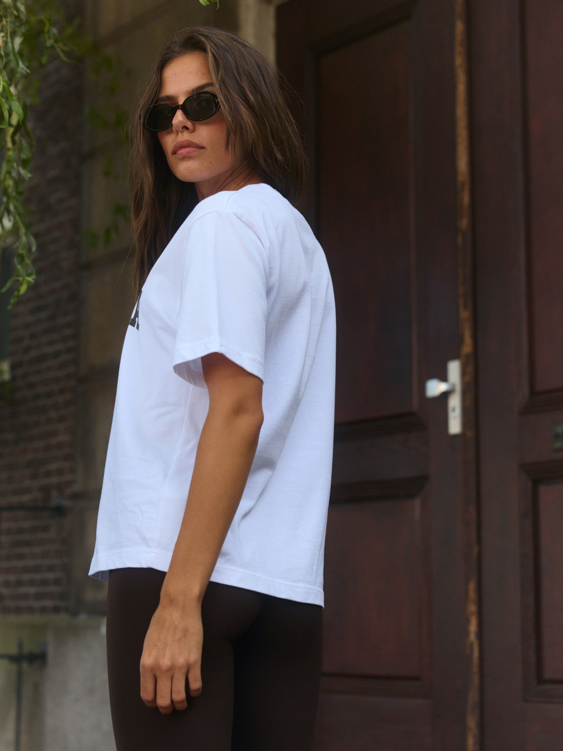 Woman wearing a white relaxed t-shirt and sunglasses standing outdoors next to a dark wooden door.