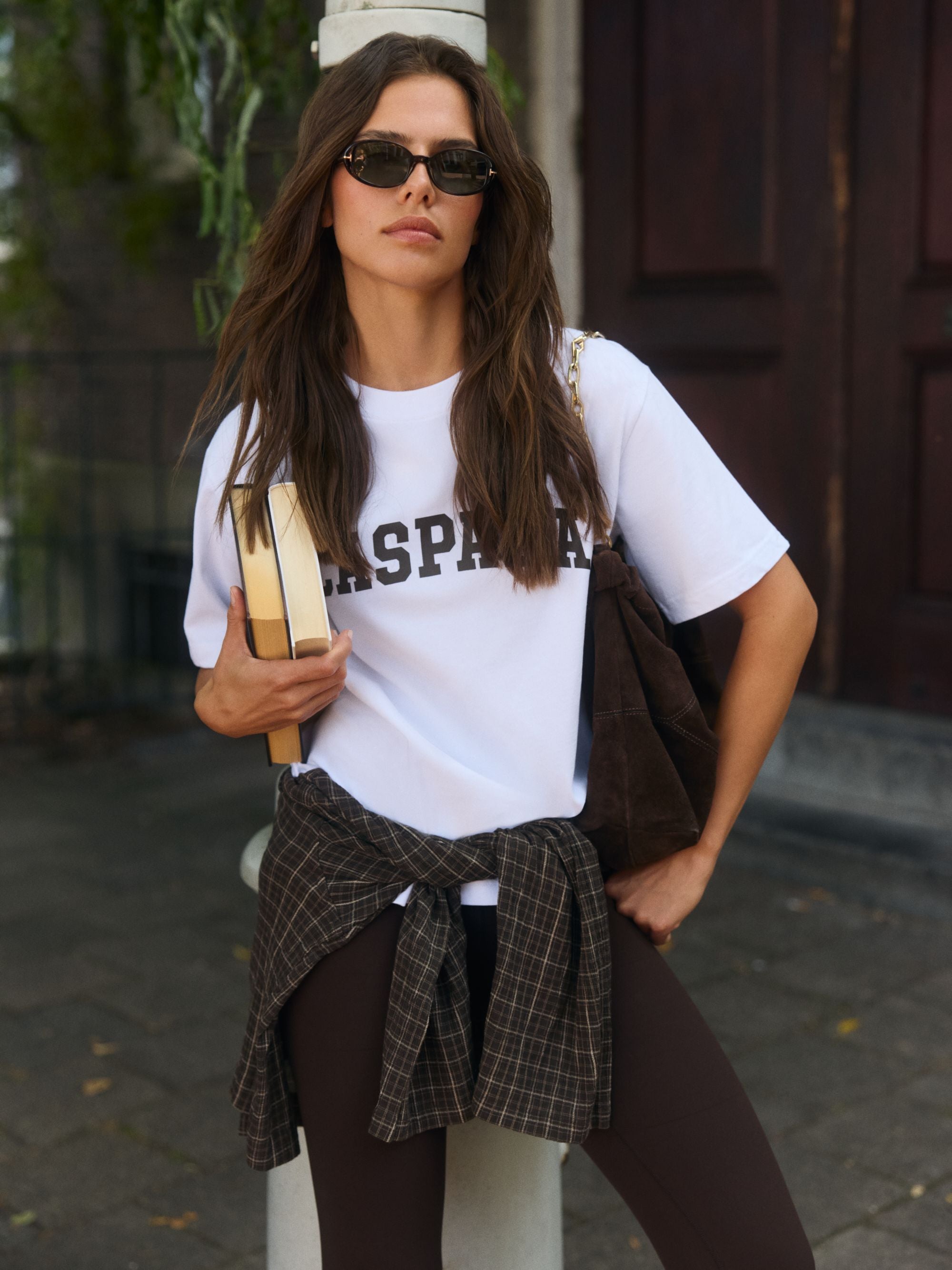 Woman wearing a white CASPARA logo t-shirt and sunglasses holding books outdoors.