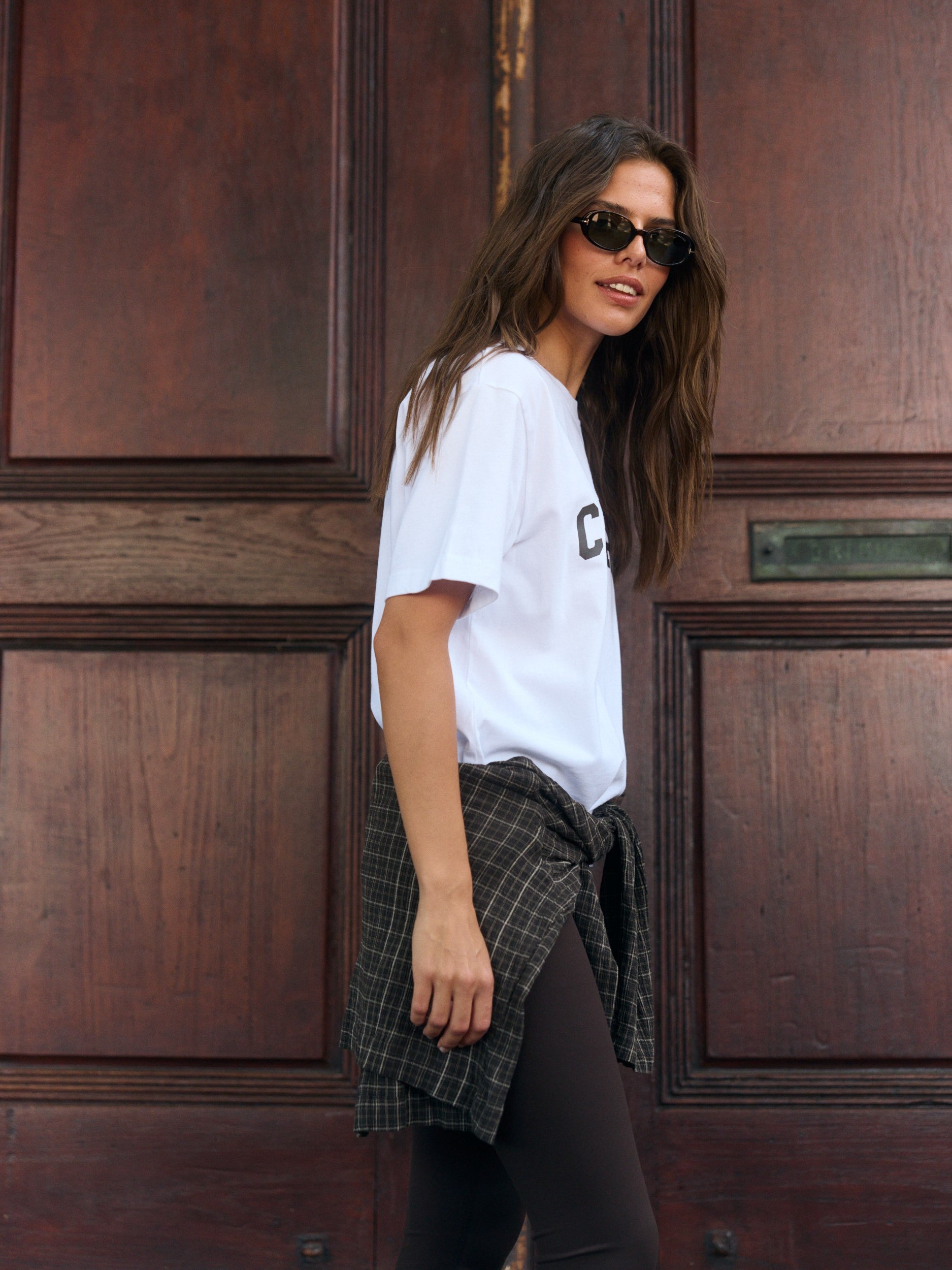 Woman wearing a white CASPARA graphic t-shirt and sunglasses in front of a wooden door.