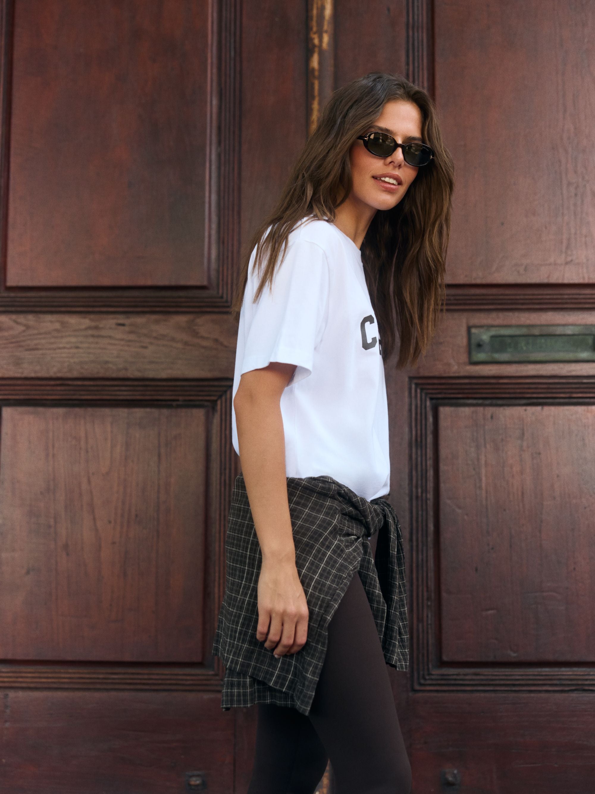 Woman wearing a white CASPARA logo t-shirt and sunglasses standing in front of a dark wooden door.