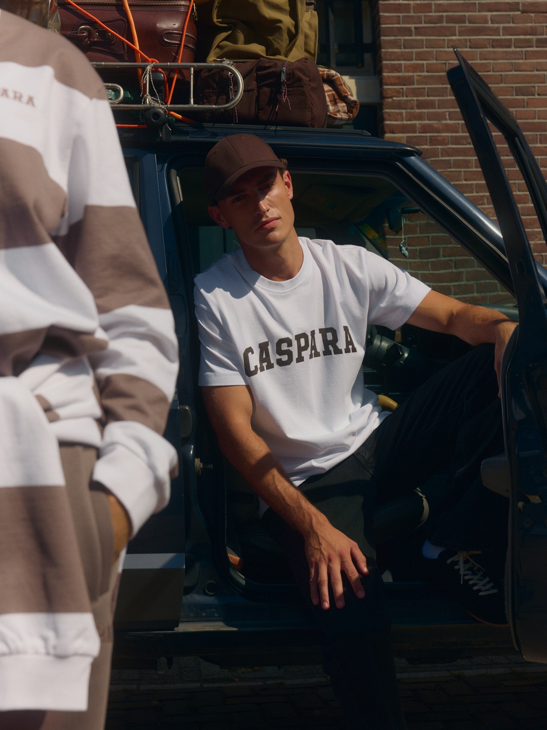 Man wearing a white CASPARA graphic t-shirt and brown cap sitting in a car packed with luggage.