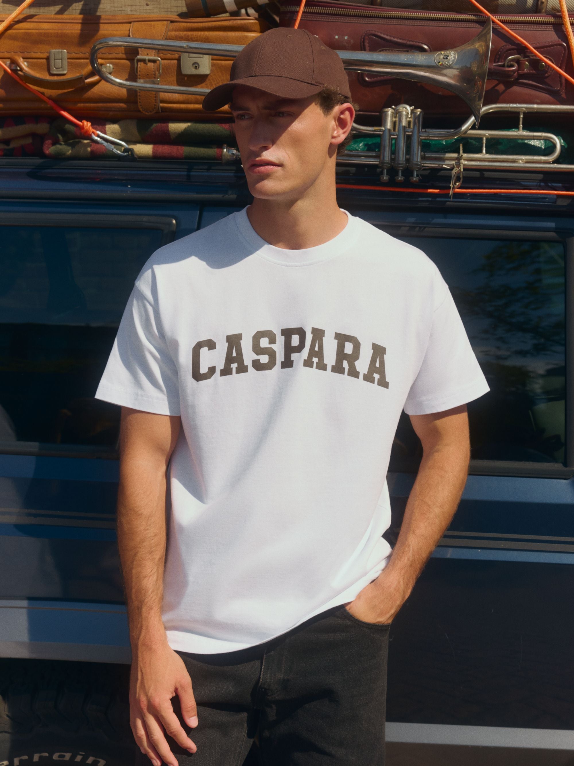 Man wearing a white CASPARA logo t-shirt and brown cap standing next to a car.
