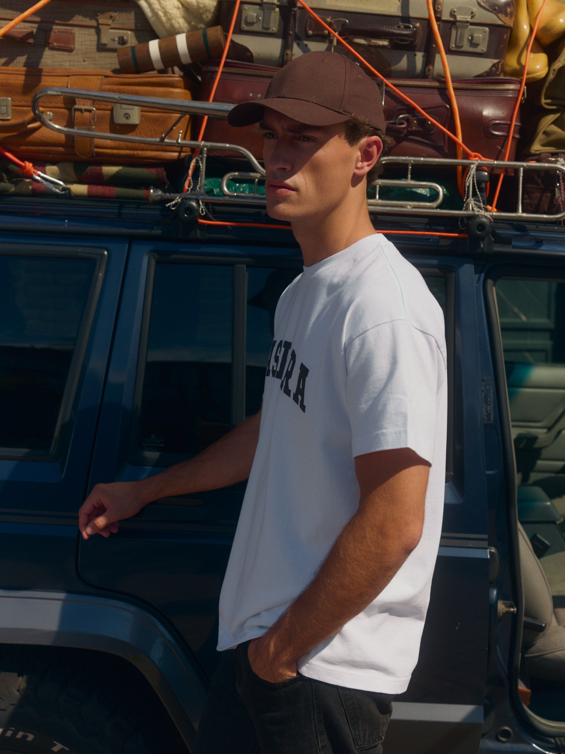 Man wearing a white oversized graphic t-shirt and brown cap beside a vintage car with luggage.