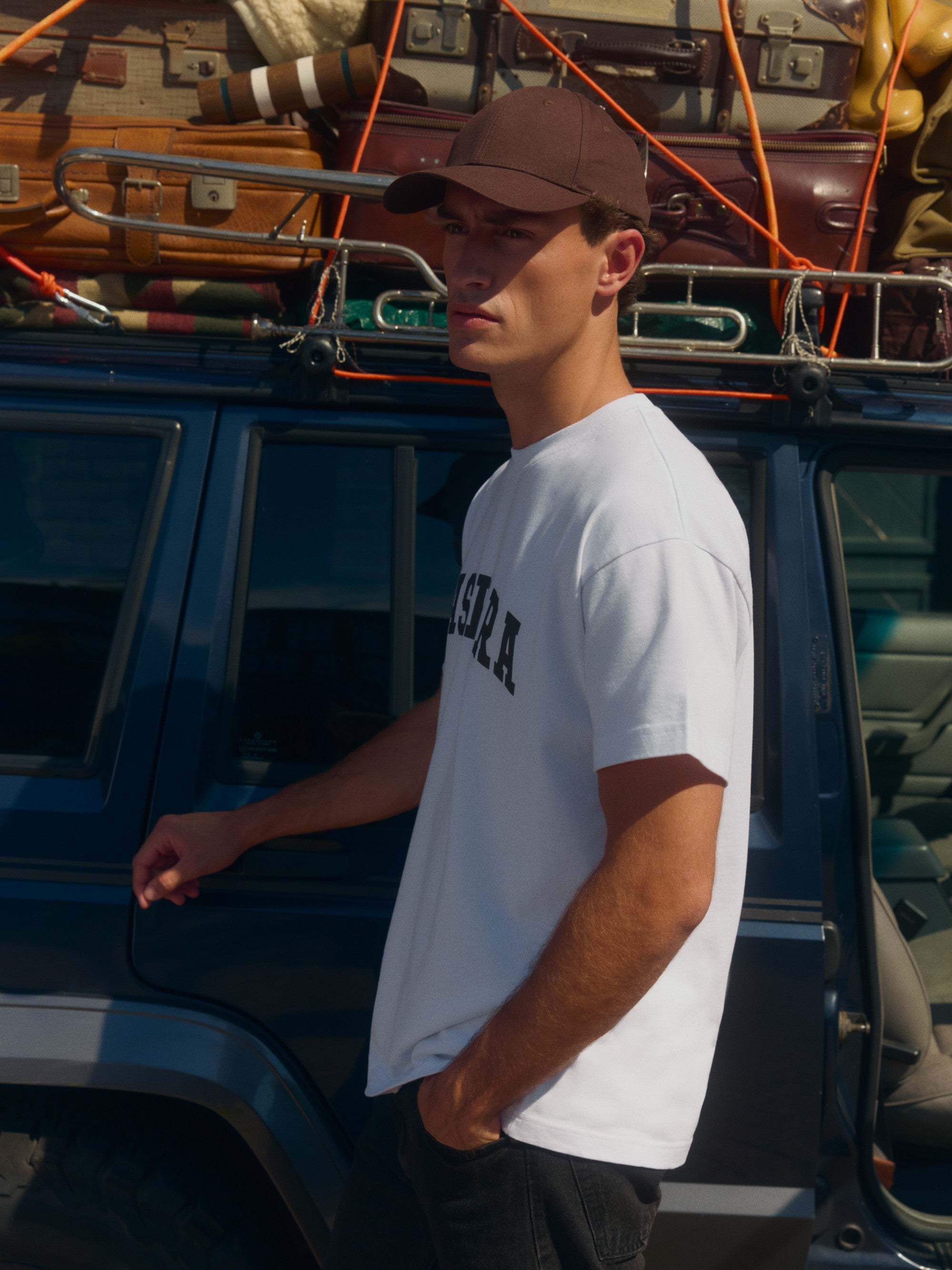 Man wearing a white CASPARA signature t-shirt and brown cap standing next to a car with luggage.
