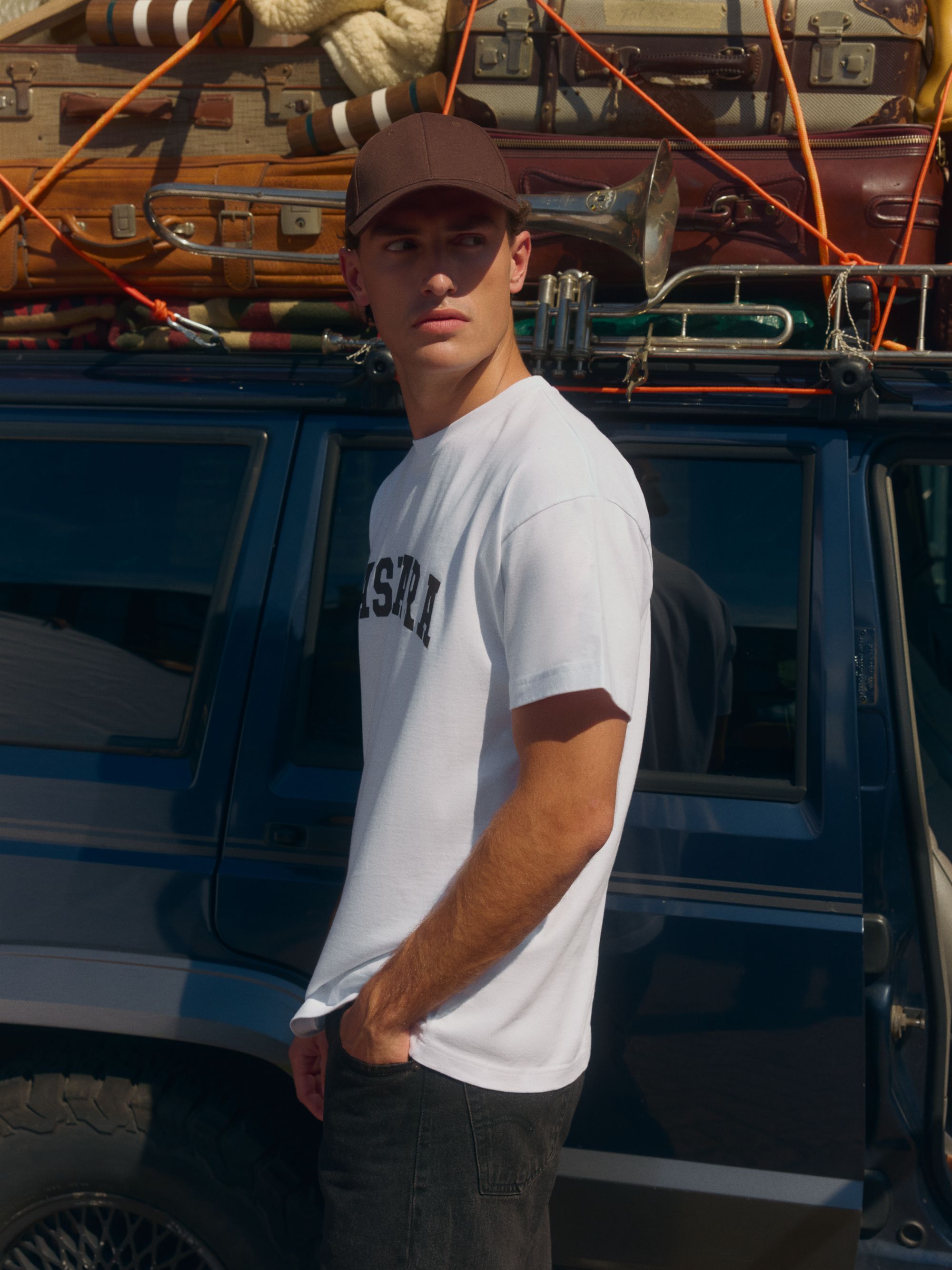 Man wearing a white CASPARA logo t-shirt and brown cap standing next to a vintage car.