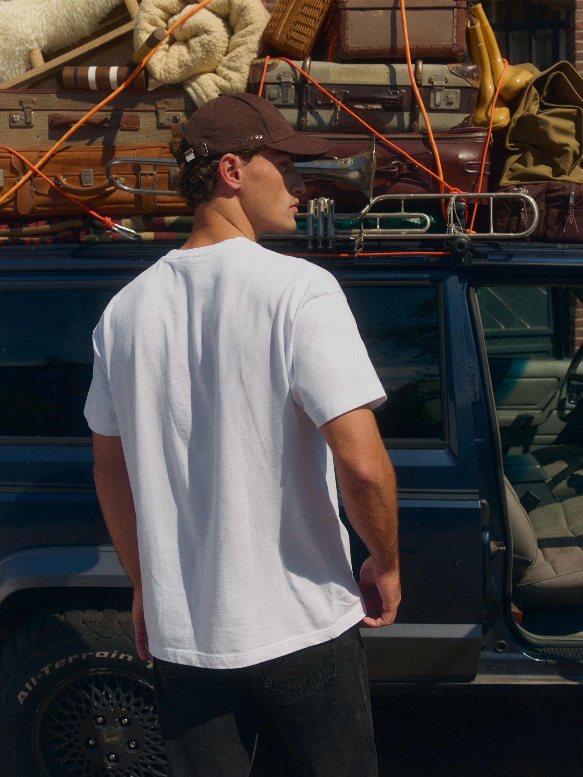Back view of a man wearing a white oversized cotton t-shirt standing next to an SUV with luggage.