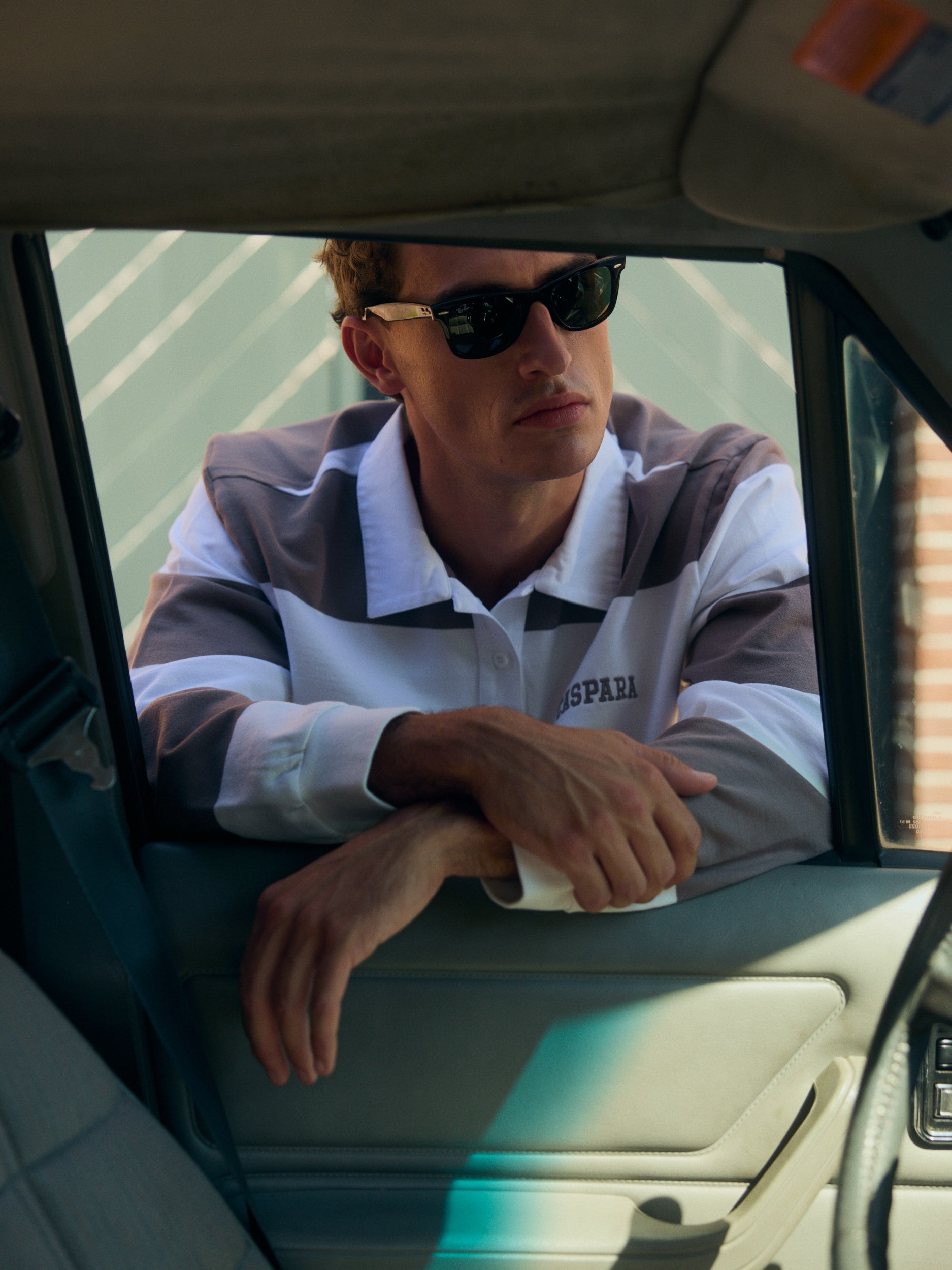 Man wearing a walnut and white striped rugby shirt and sunglasses leaning on an open car window.