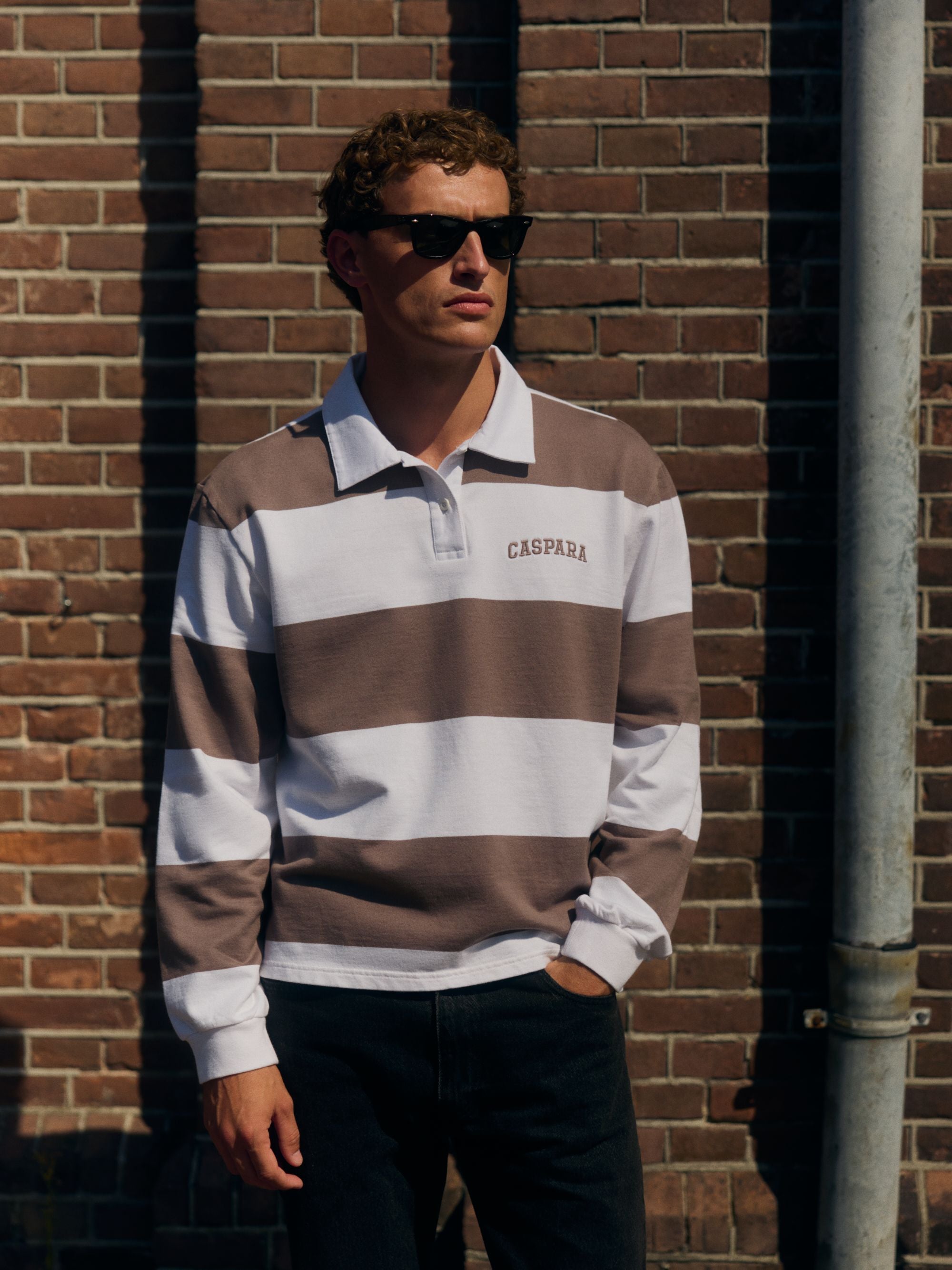 Man wearing a walnut and white striped CASPARA logo rugby shirt and sunglasses against a brick wall.