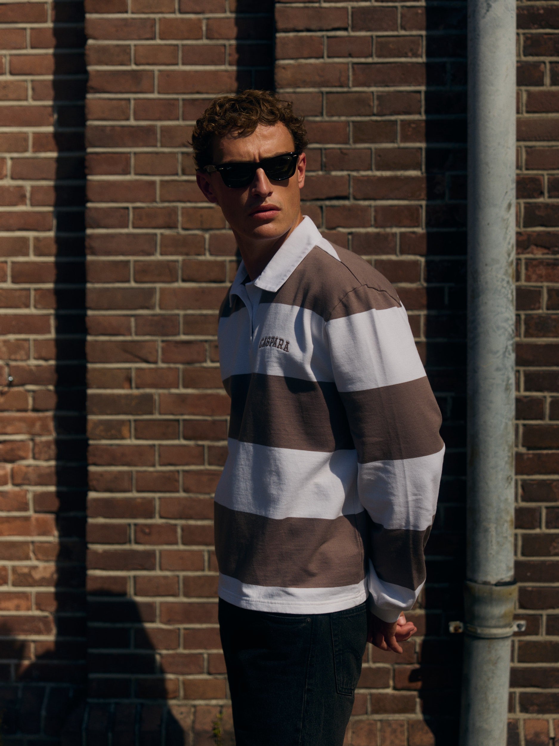 Man wearing a walnut and white striped rugby shirt and sunglasses against a brick wall.