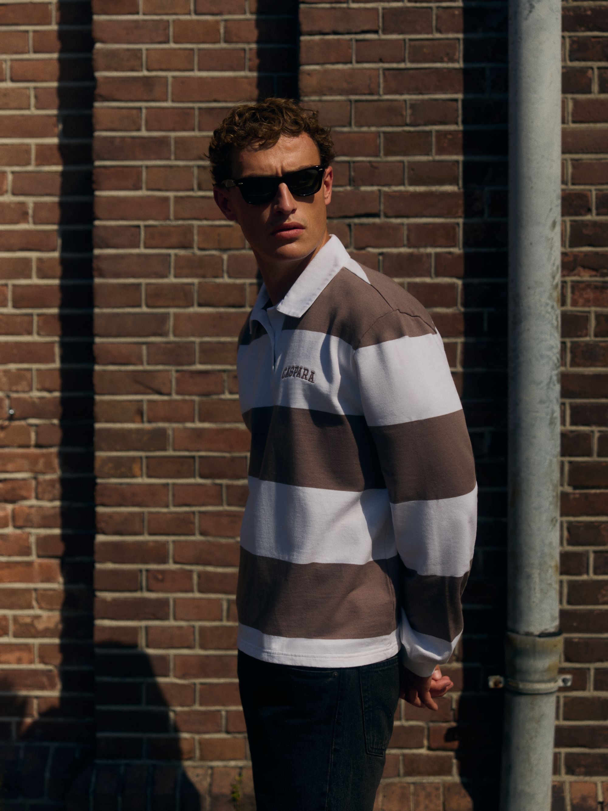 Man wearing a walnut and white striped CASPARA rugby shirt and sunglasses against a brick wall.