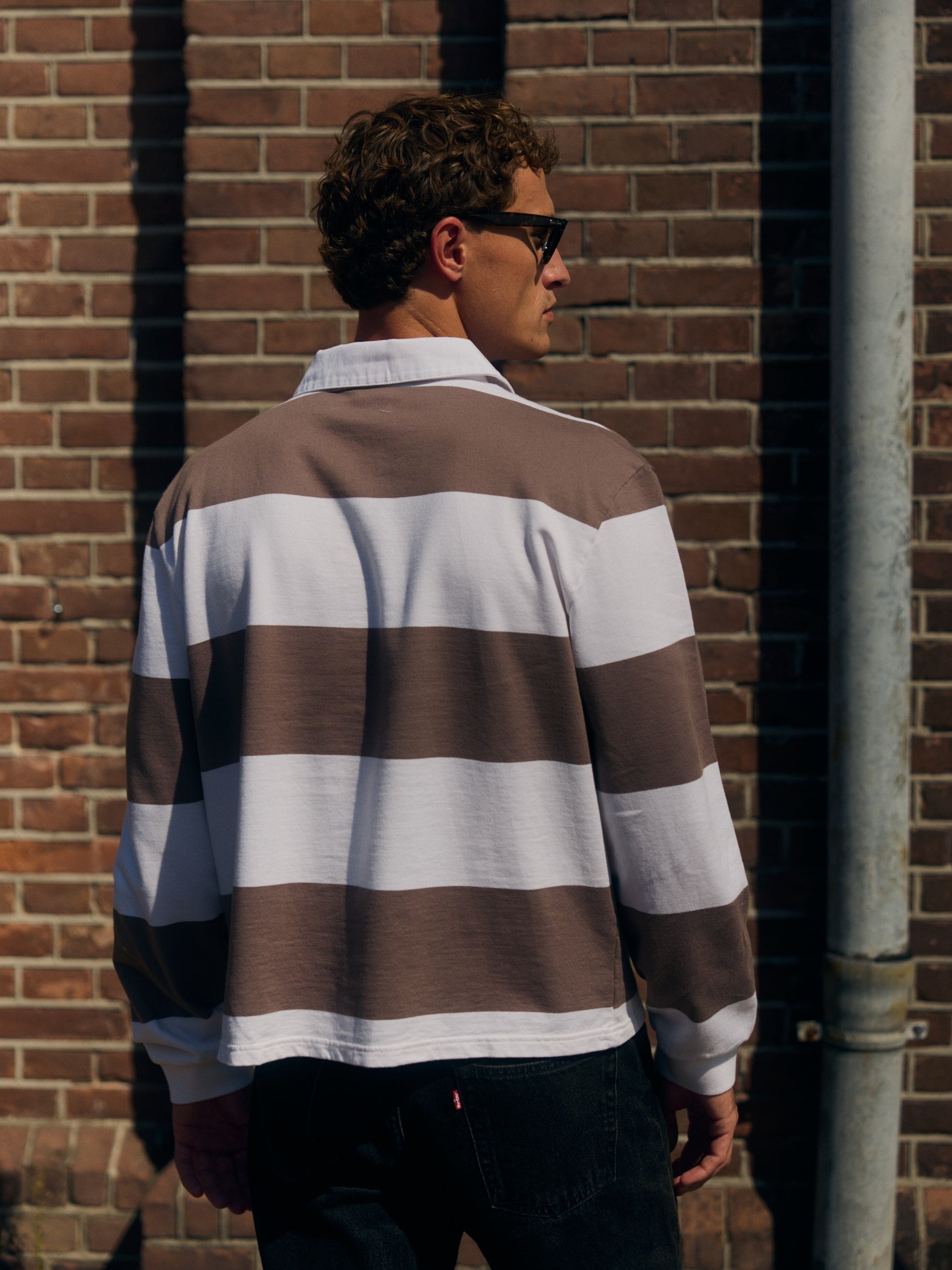 Back view of a man wearing a walnut and white striped rugby shirt against a brick wall.