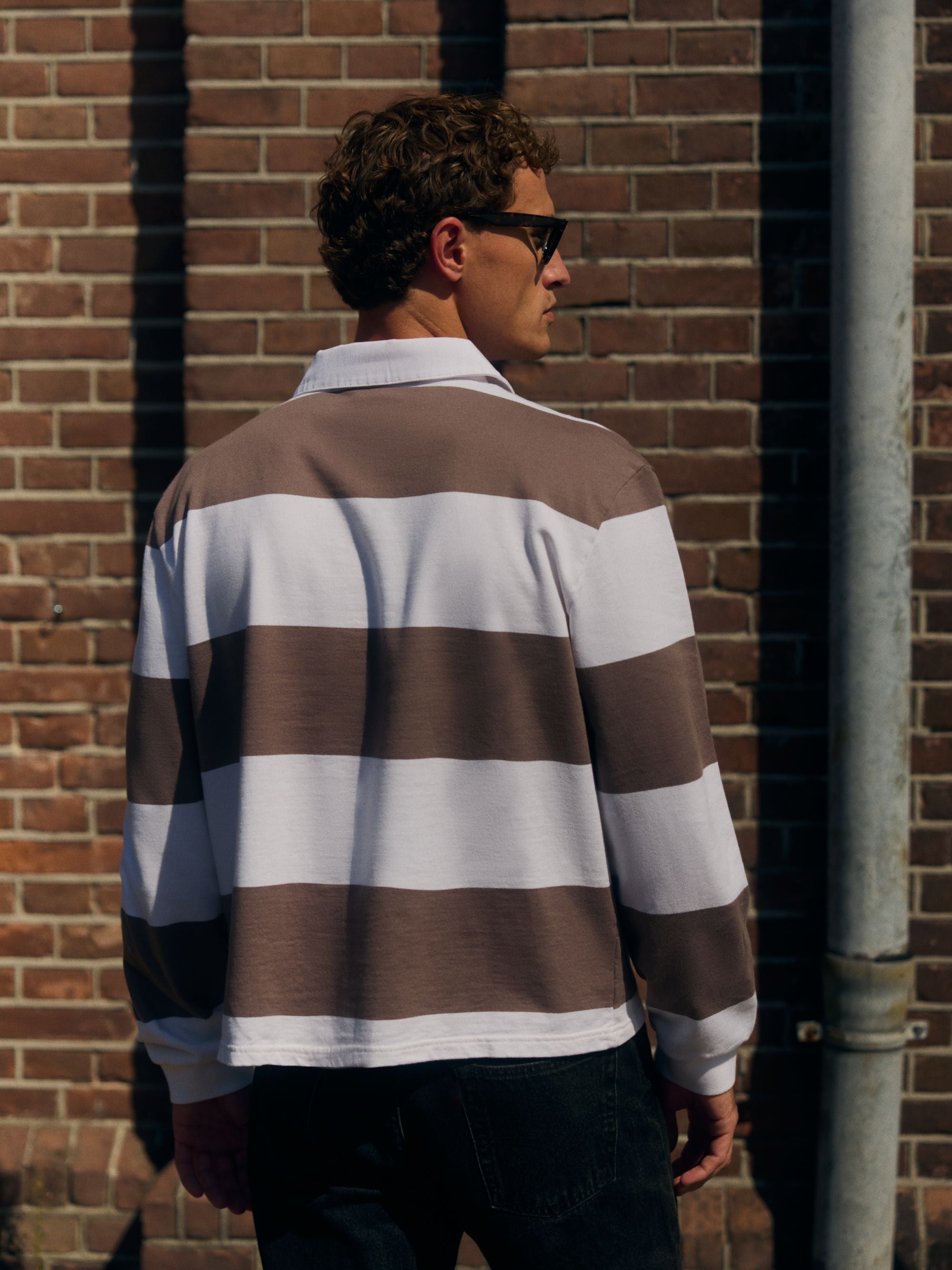 Back view of a man wearing a walnut and white striped CASPARA rugby shirt against a brick wall.