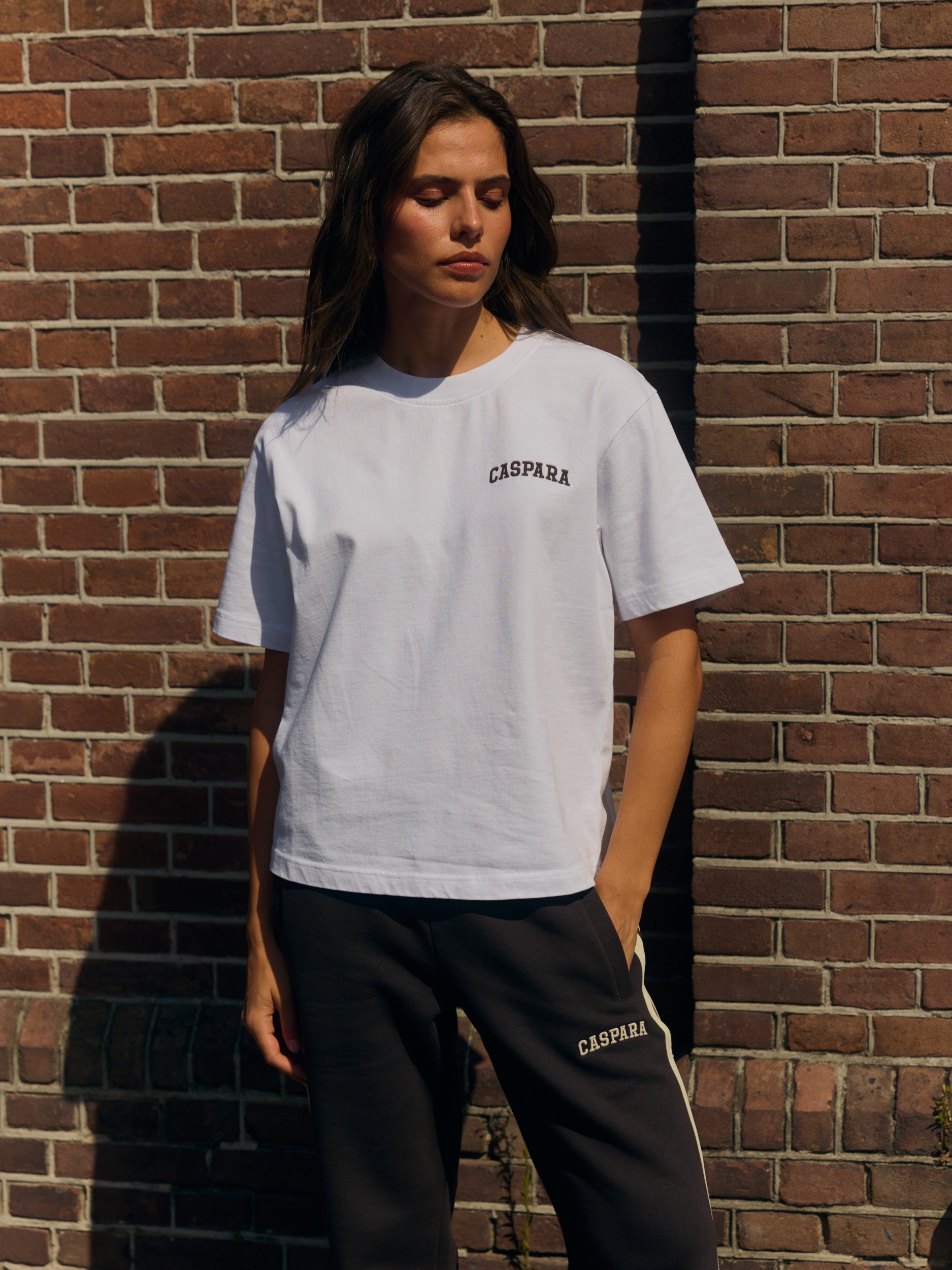 Woman wearing a white embroidered logo t-shirt and espresso sweatpants against a brick wall.
