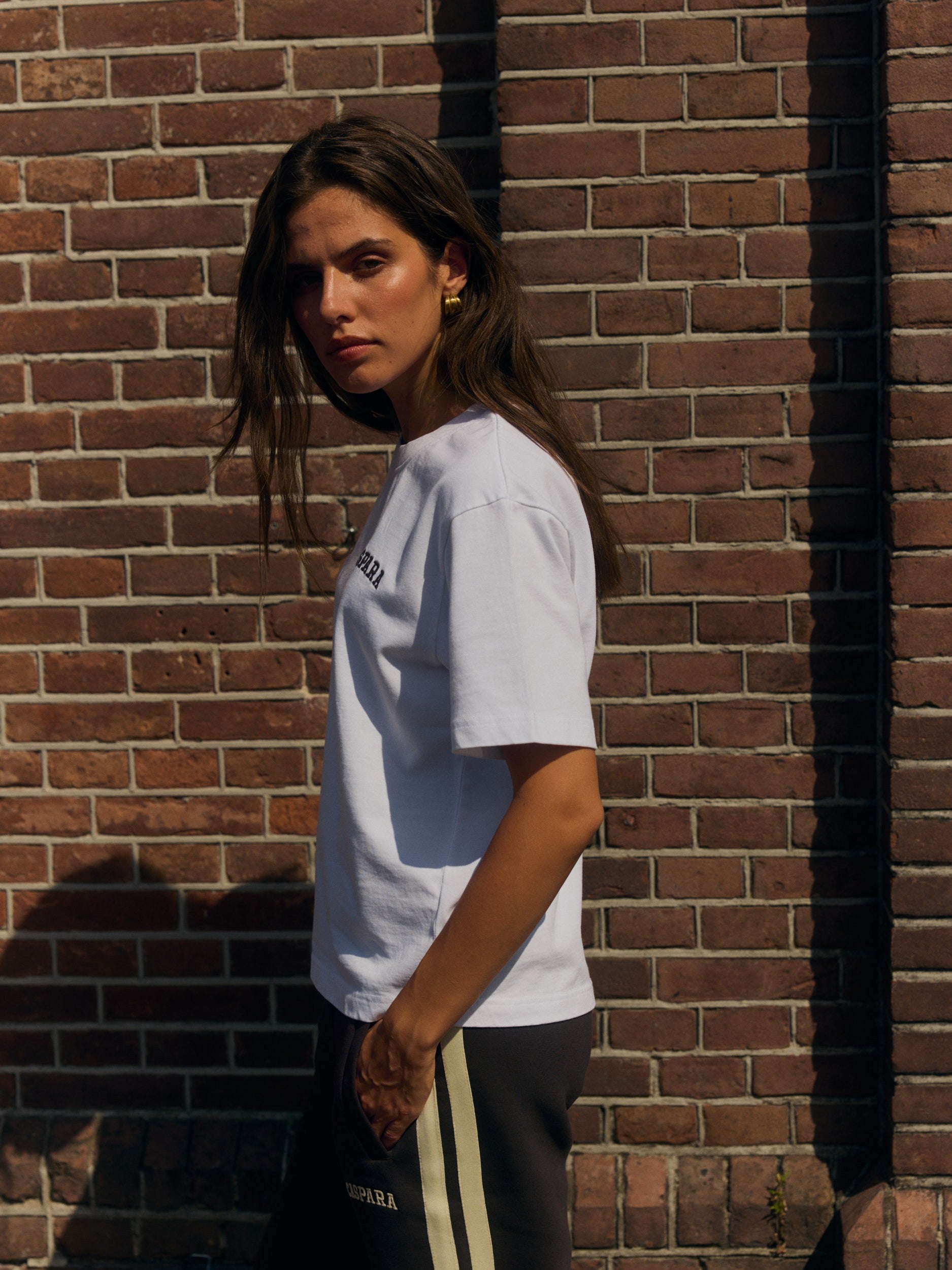 Woman wearing a white embroidered cotton t-shirt and dark sweatpants against a brick wall.
