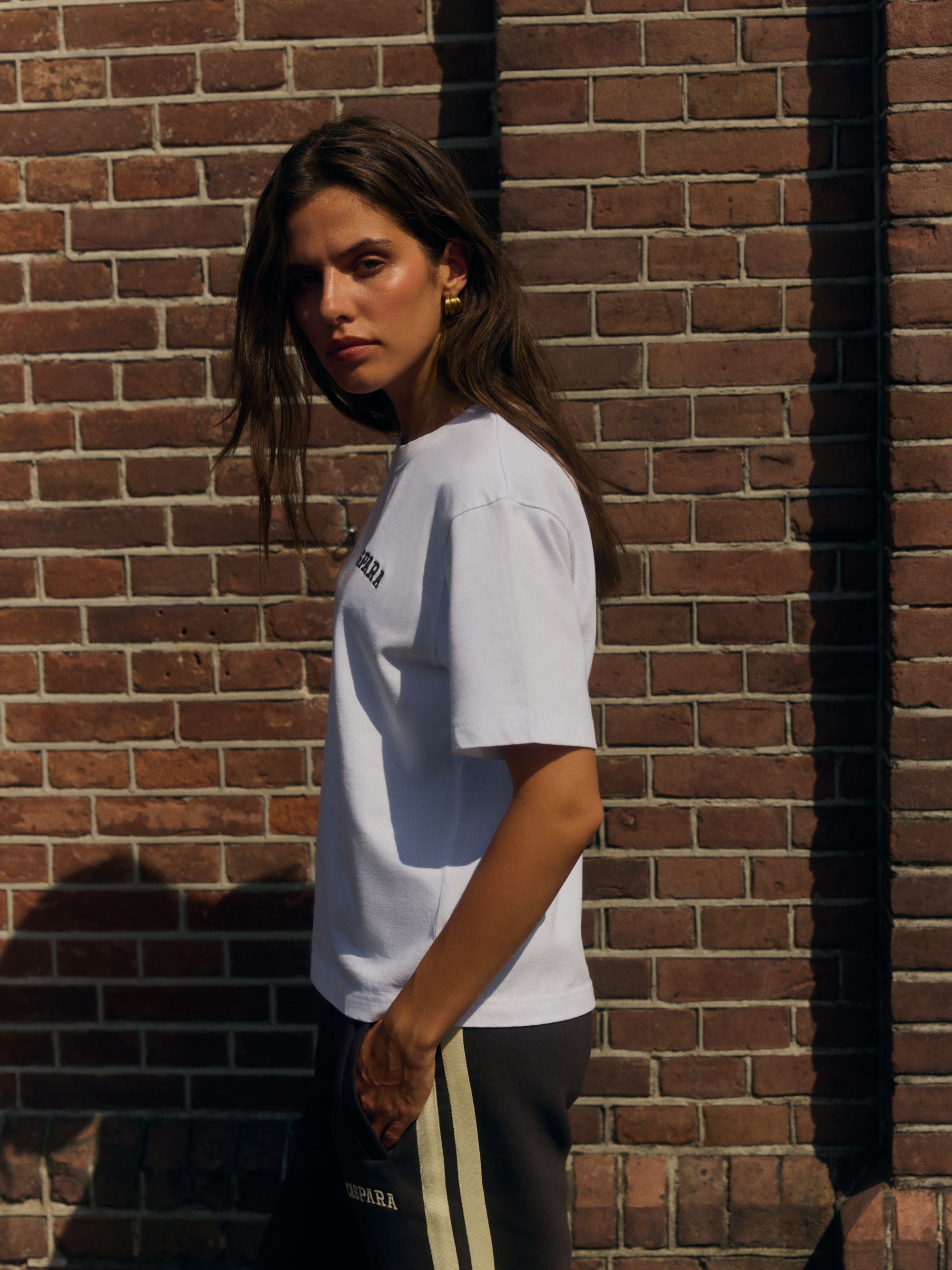 Woman wearing a white CASPARA embroidered logo t-shirt against a brick wall.
