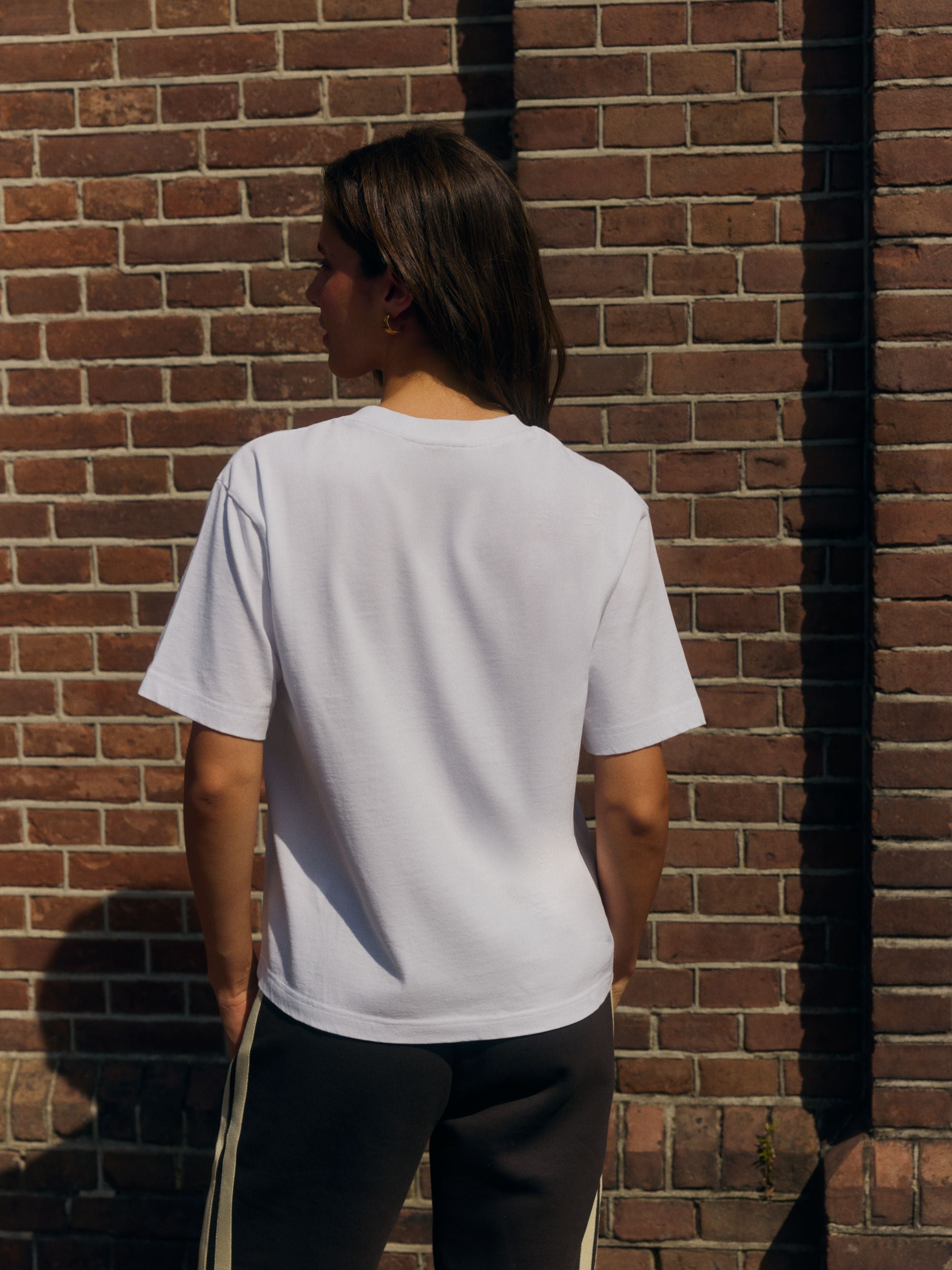Back view of a woman wearing a white CASPARA cotton t-shirt against a brick wall.