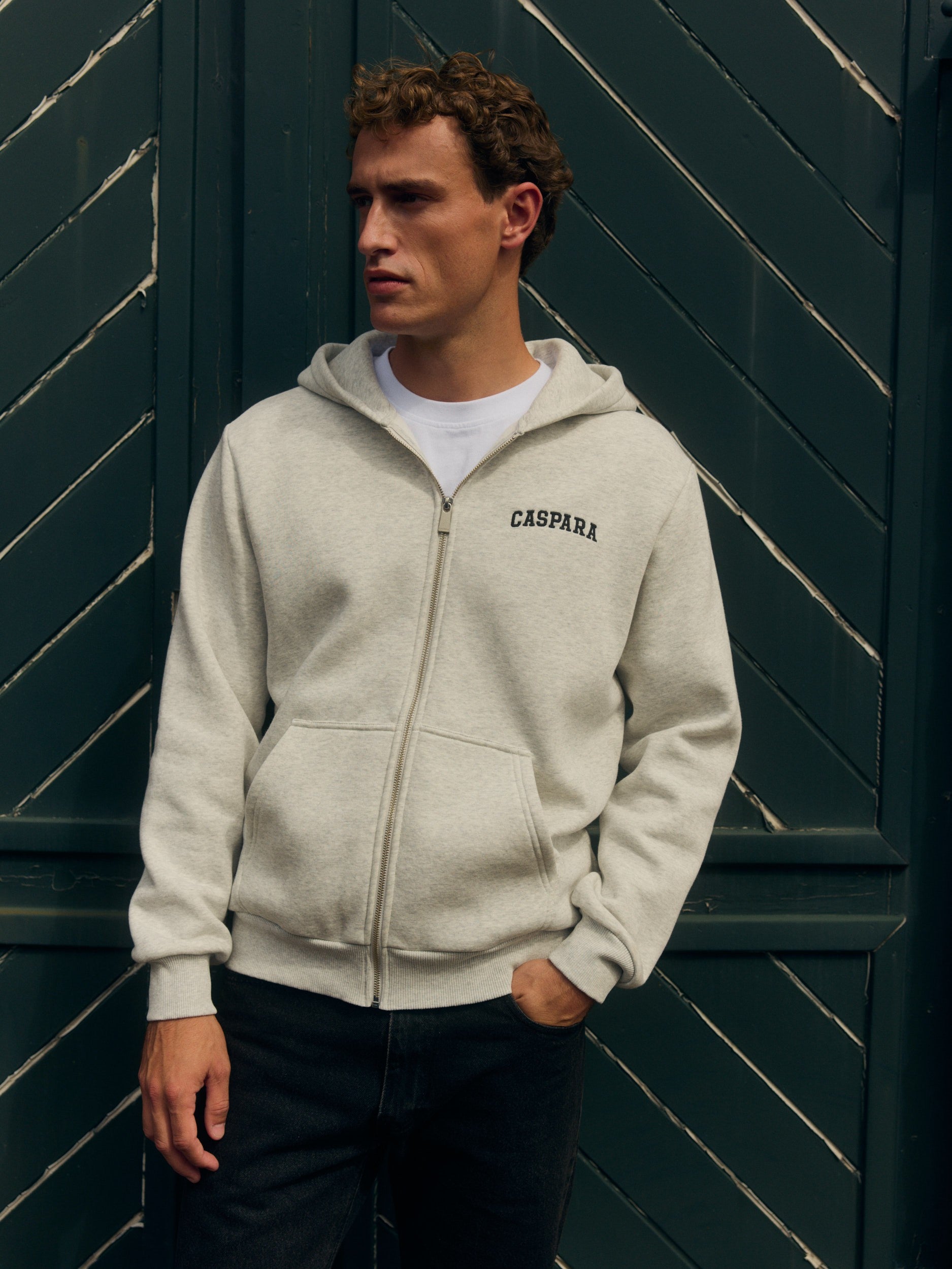 Man wearing a grey melange zip hoodie standing against a dark green wooden door.