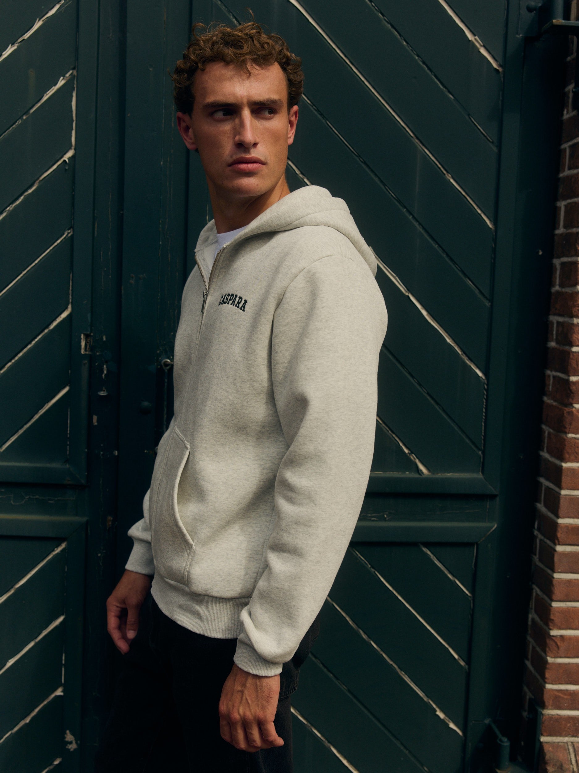 Man wearing a grey melange zip hoodie standing against a dark green wooden door.
