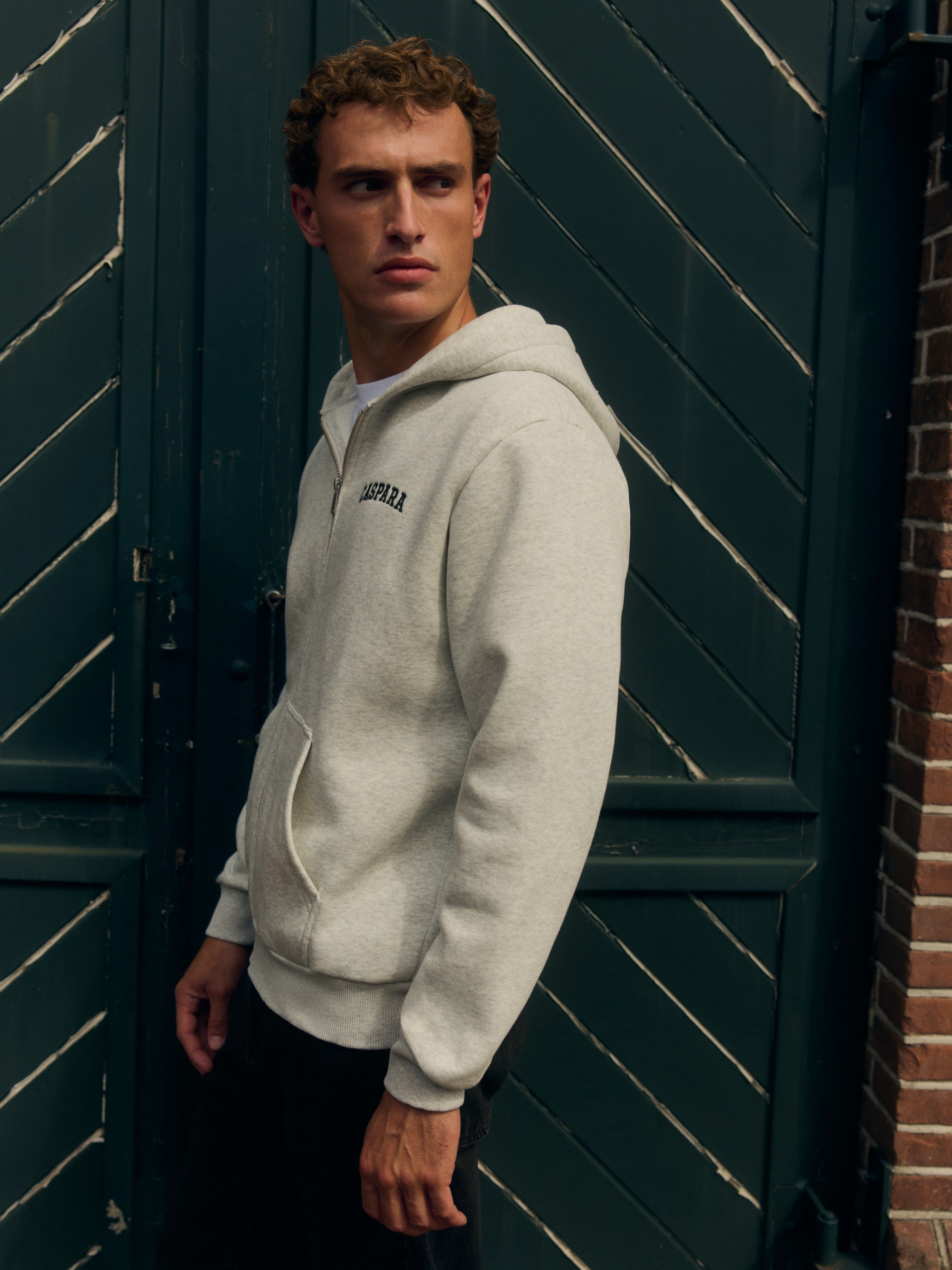 Man wearing a grey melange CASPARA logo zip hoodie in front of a dark green door.