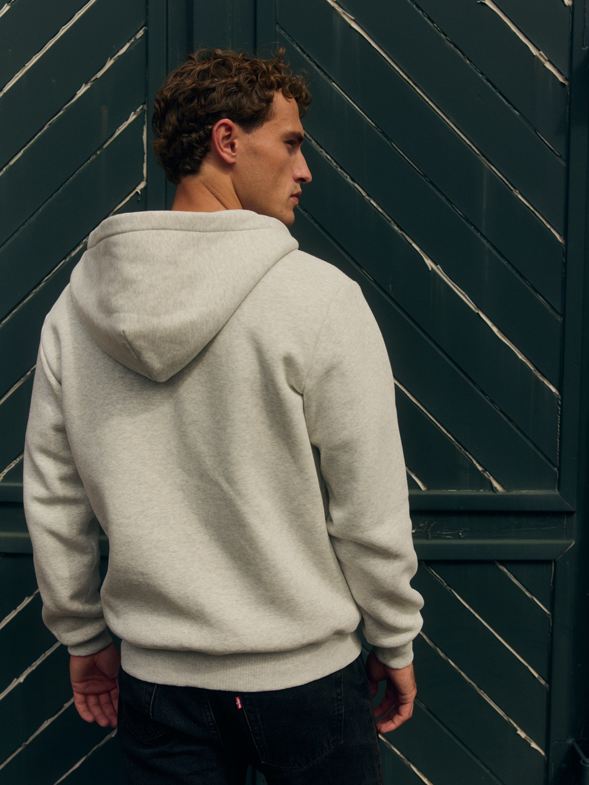 Back view of a man wearing a grey melange zip hoodie against a dark green wooden door.
