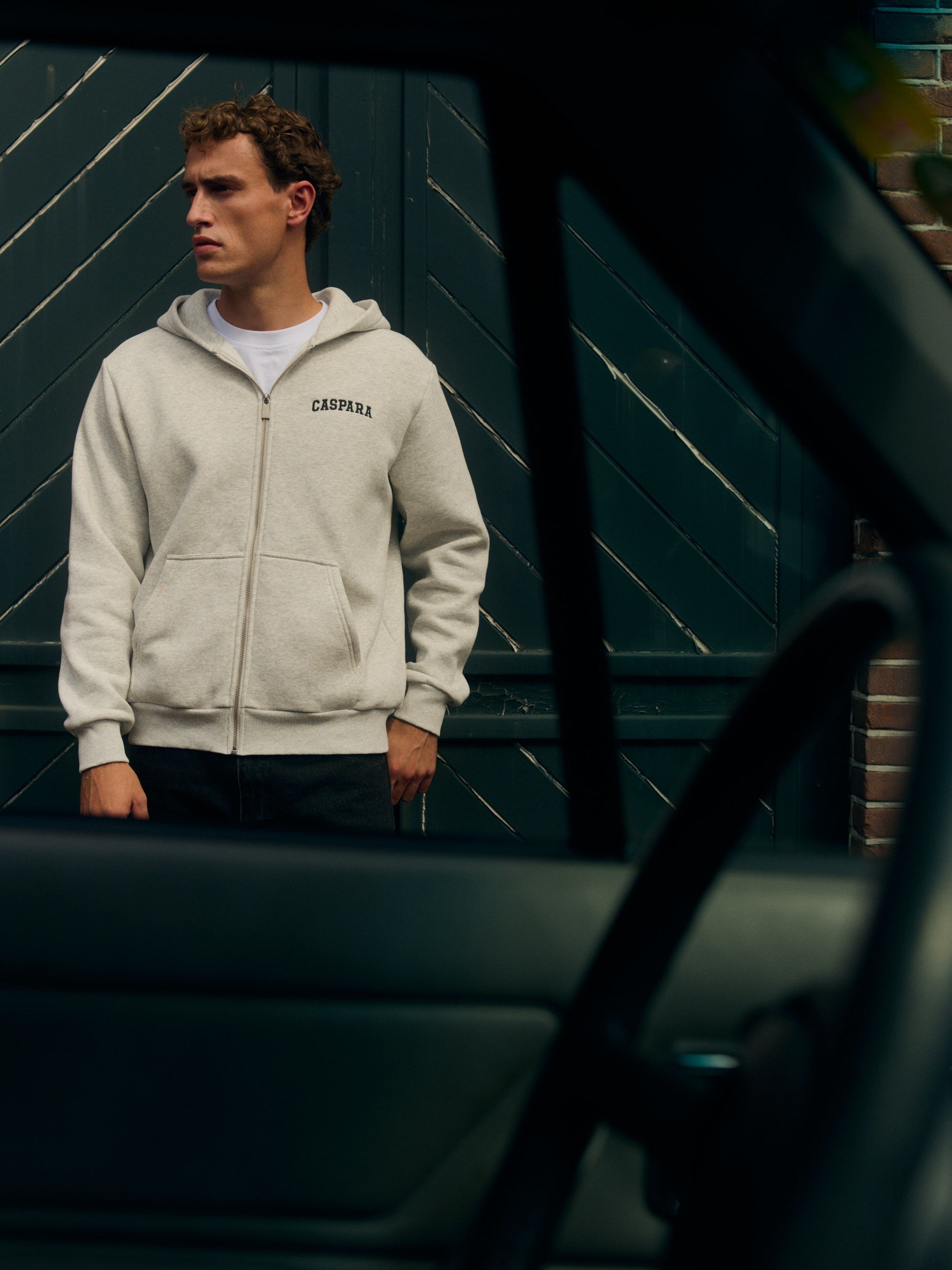 Man wearing a grey melange zip hoodie standing outside in front of dark green wooden doors.