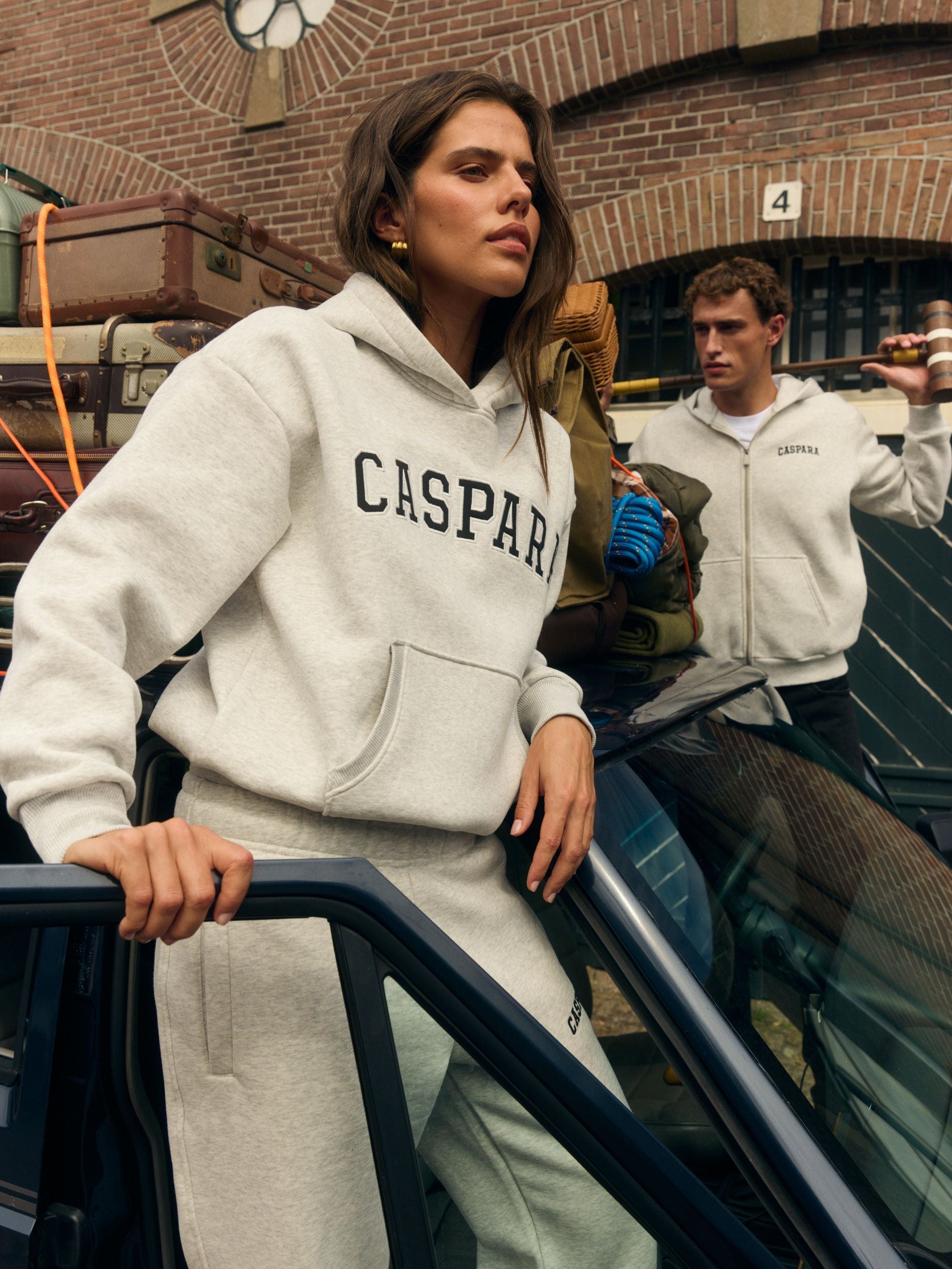 Woman and man wearing grey CASPARA hoodies beside a vintage car packed with luggage.