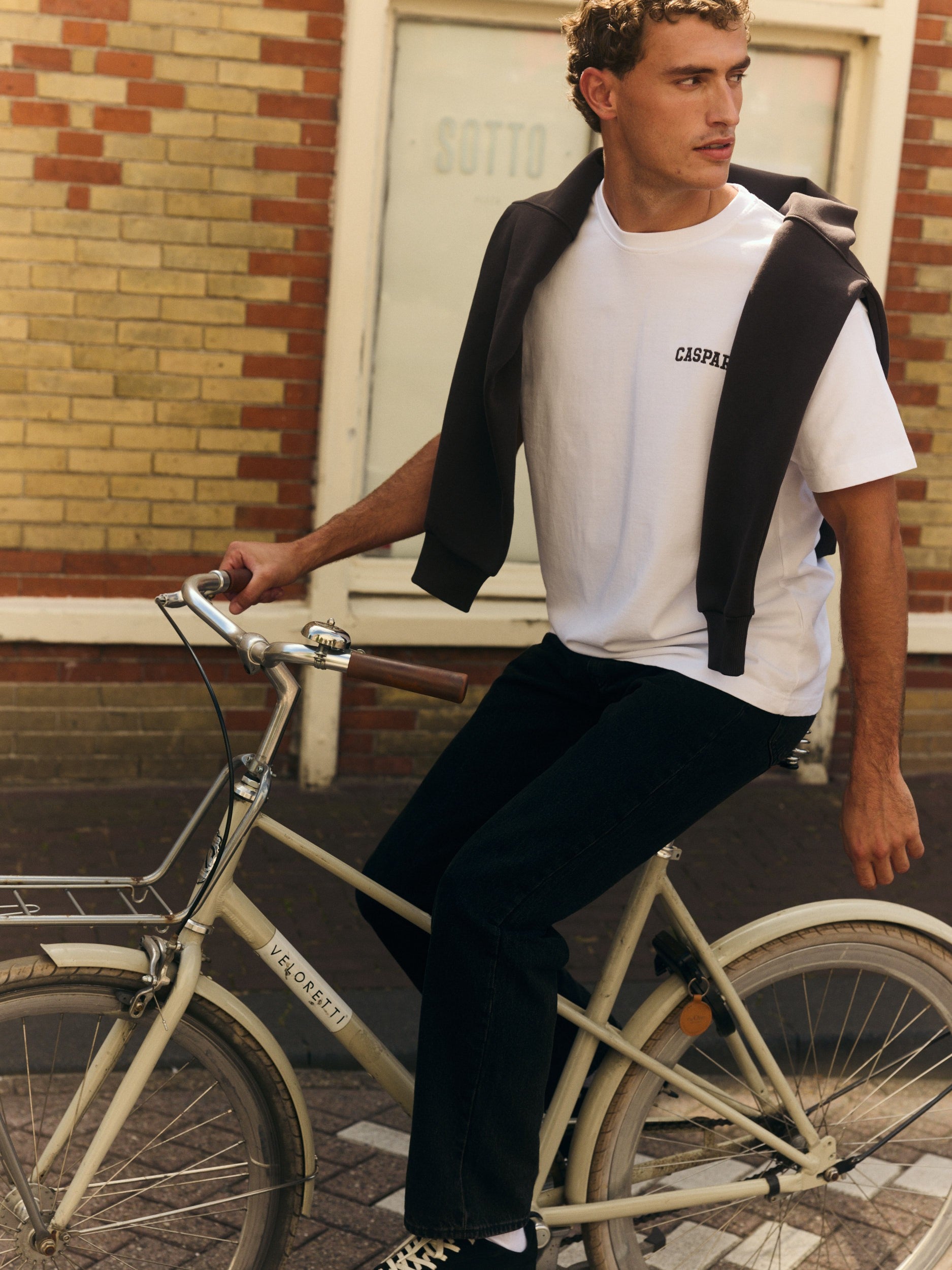 Man wearing a white embroidered t-shirt and draped sweater riding a bicycle on a city street.