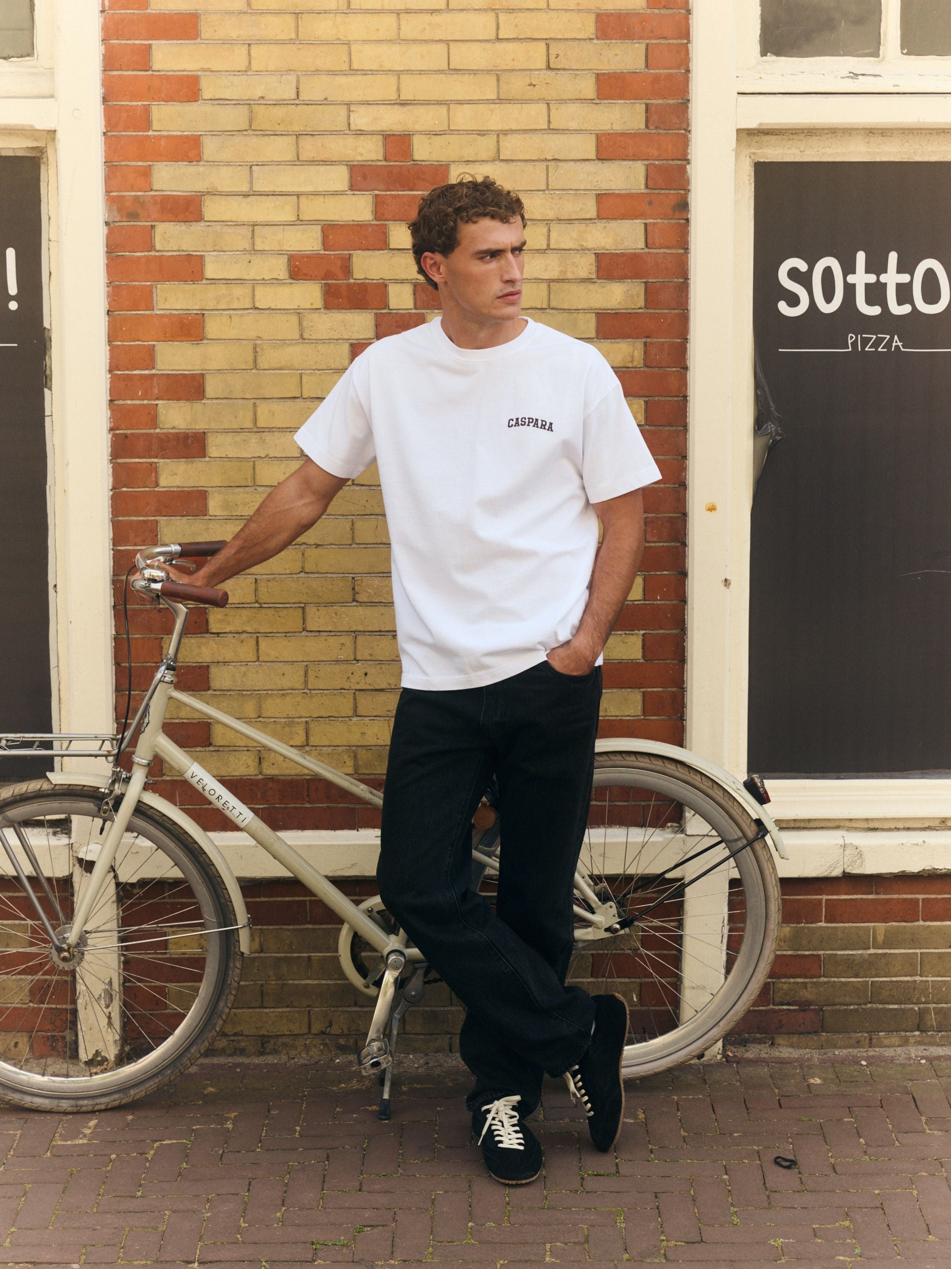 Man wearing a white embroidered logo t-shirt and black jeans leaning on a bicycle against a brick wall.