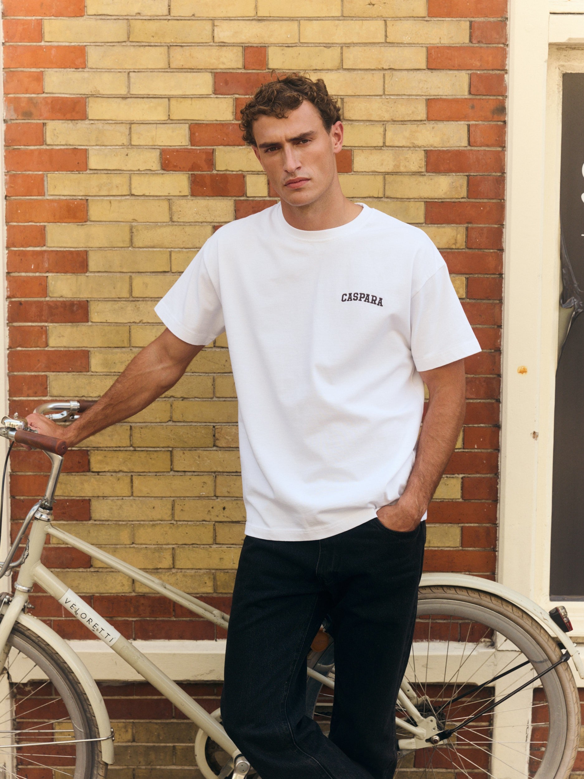 Man wearing a white embroidered t-shirt standing beside a bicycle against a brick wall.