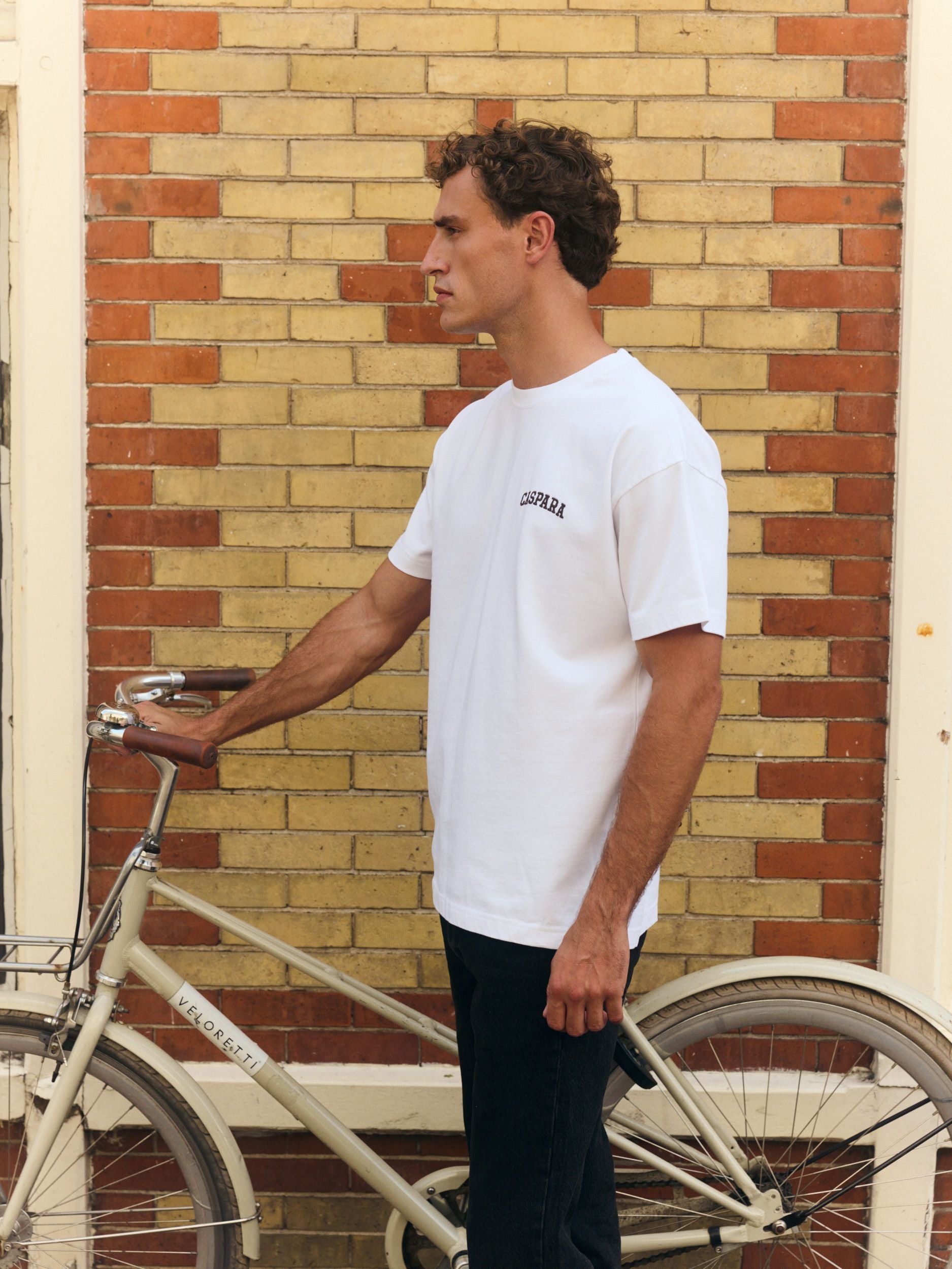 Man wearing a white embroidered cotton t-shirt standing with a bicycle against a brick wall.