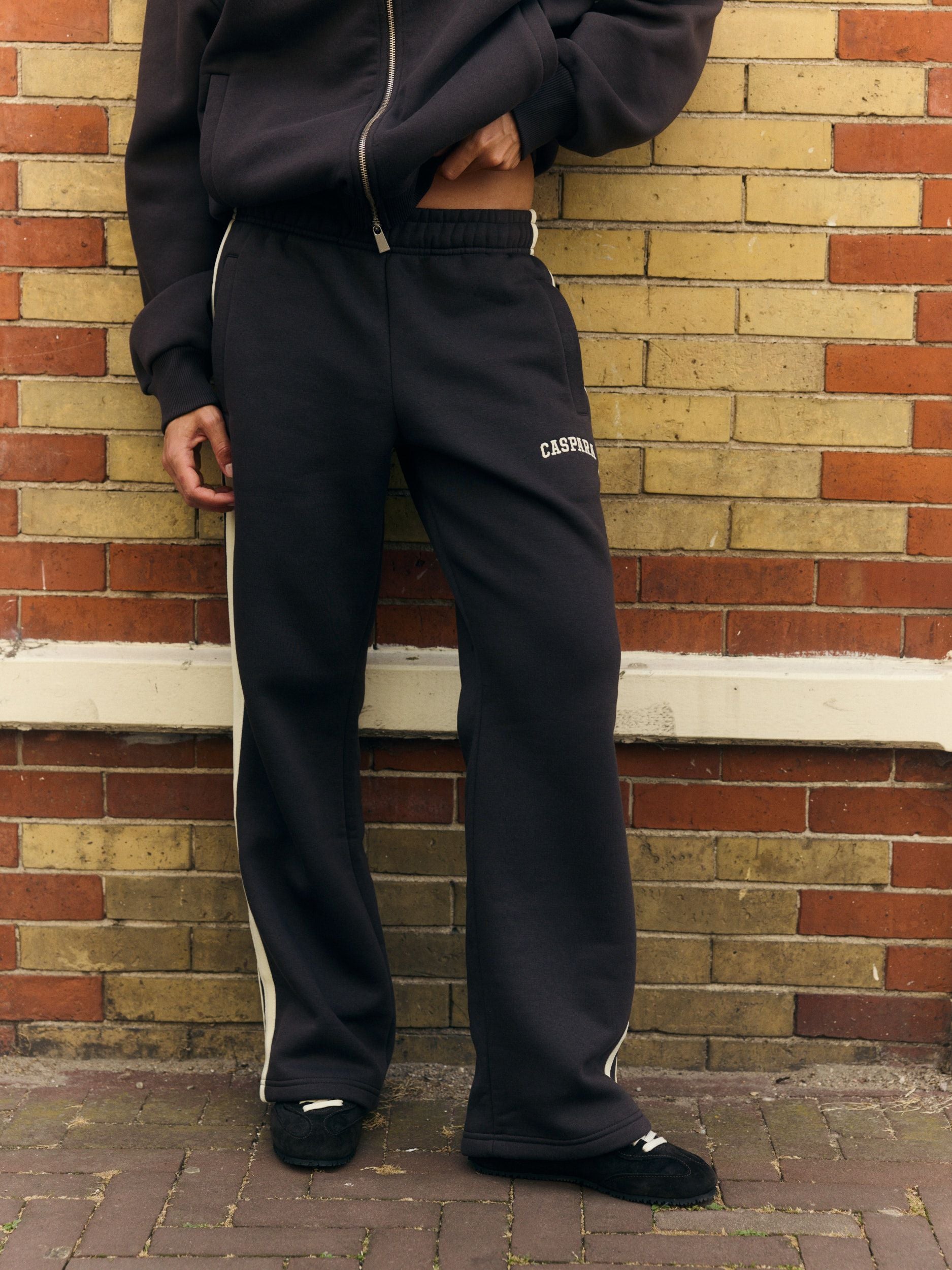 Woman wearing espresso straight-leg sweatpants with white side piping against a brick wall.