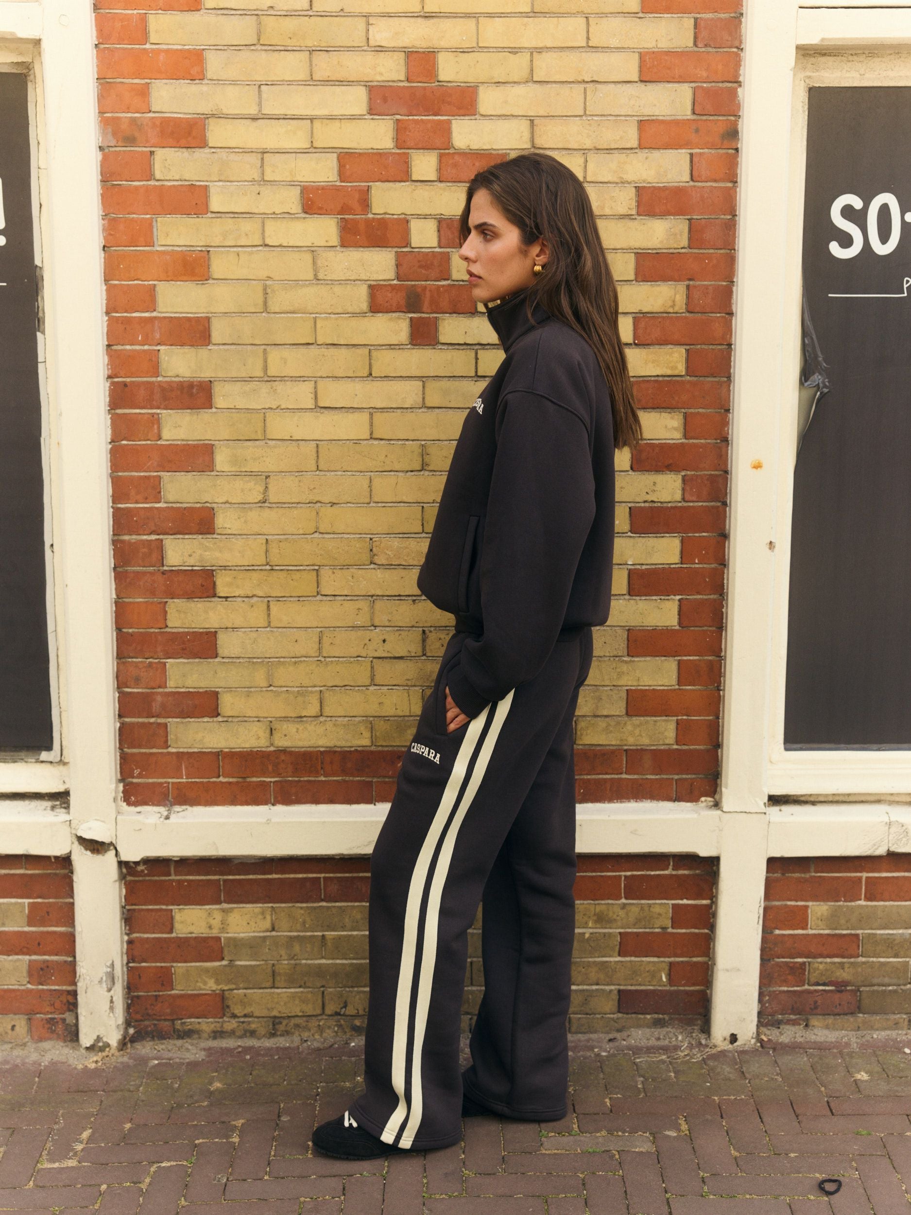 Woman wearing espresso straight-leg sweatpants with side stripes and matching sweatshirt against a brick wall.