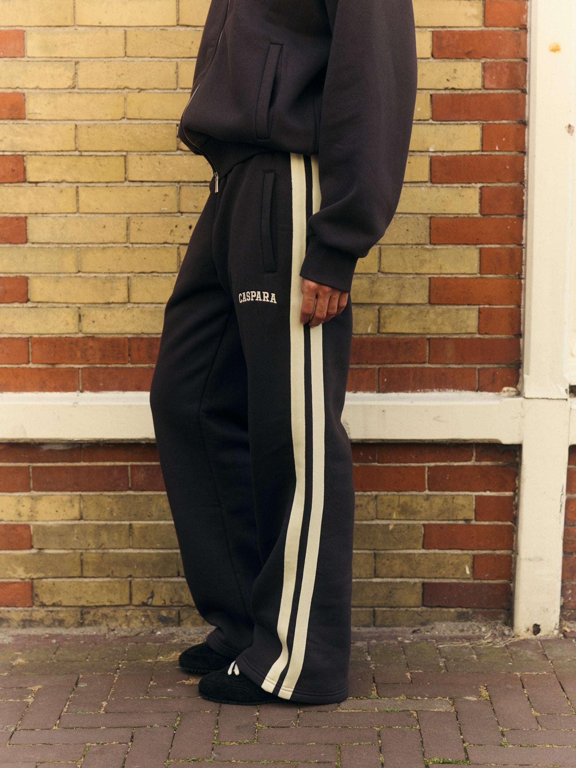 Woman wearing espresso straight-leg sweatpants with cream side stripes against a brick wall.